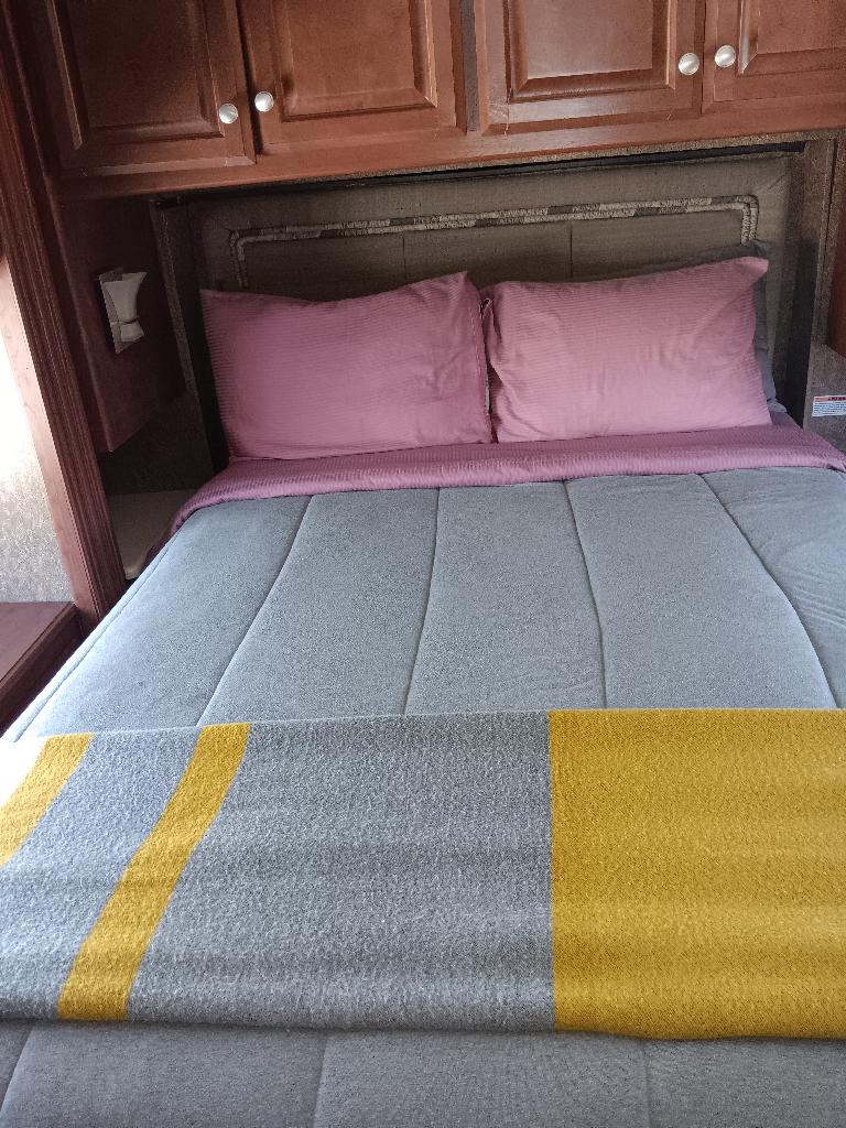 Cozy RV rental sleeping area with pink pillows, grey comforter, and a yellow-striped blanket under wooden cabinets.. Thor Palazzo 2014