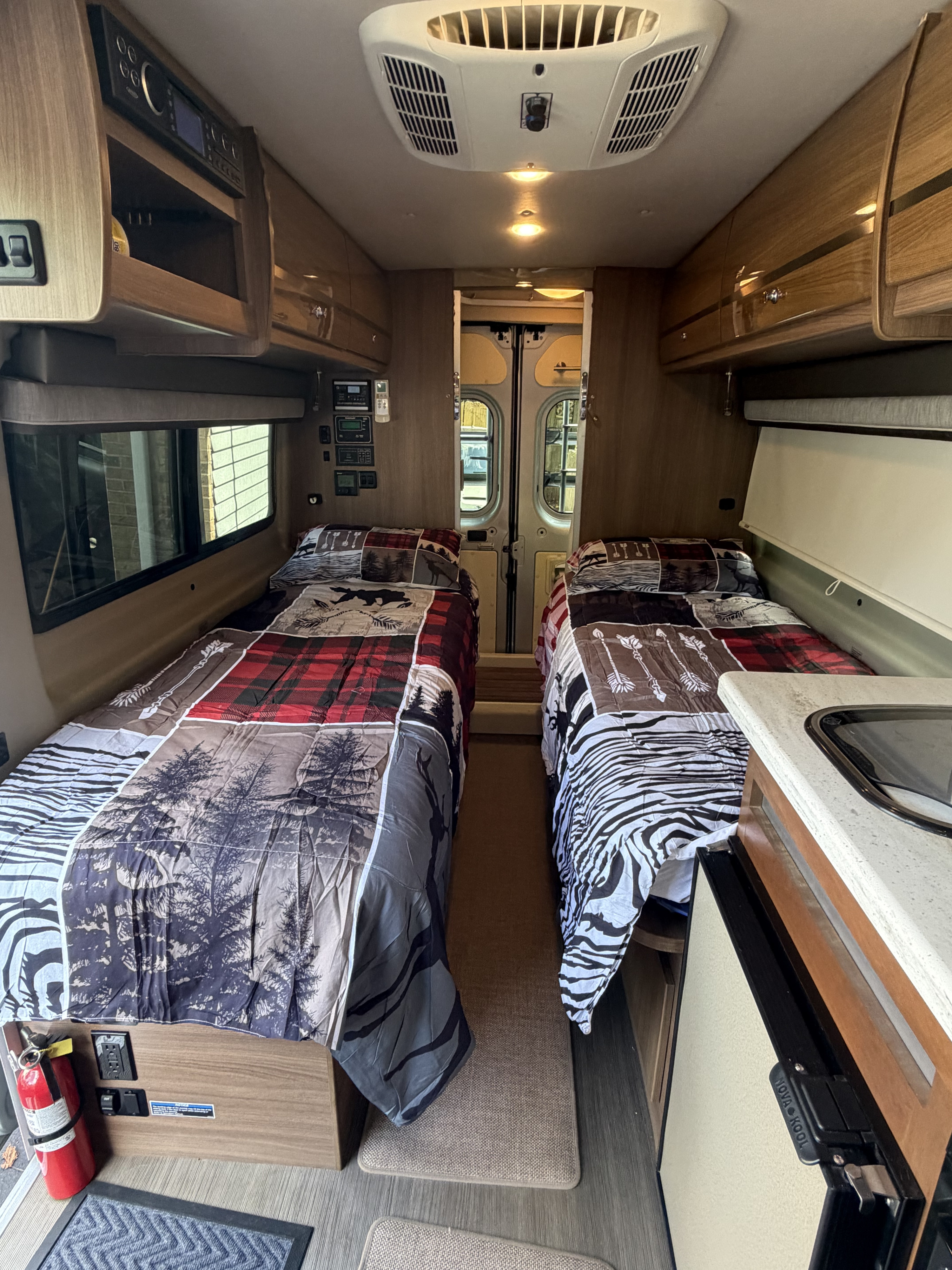 RV rental interior: two single beds with patterned comforters, overhead storage, and a compact kitchen area are visible.. Winnebago Travato 2016