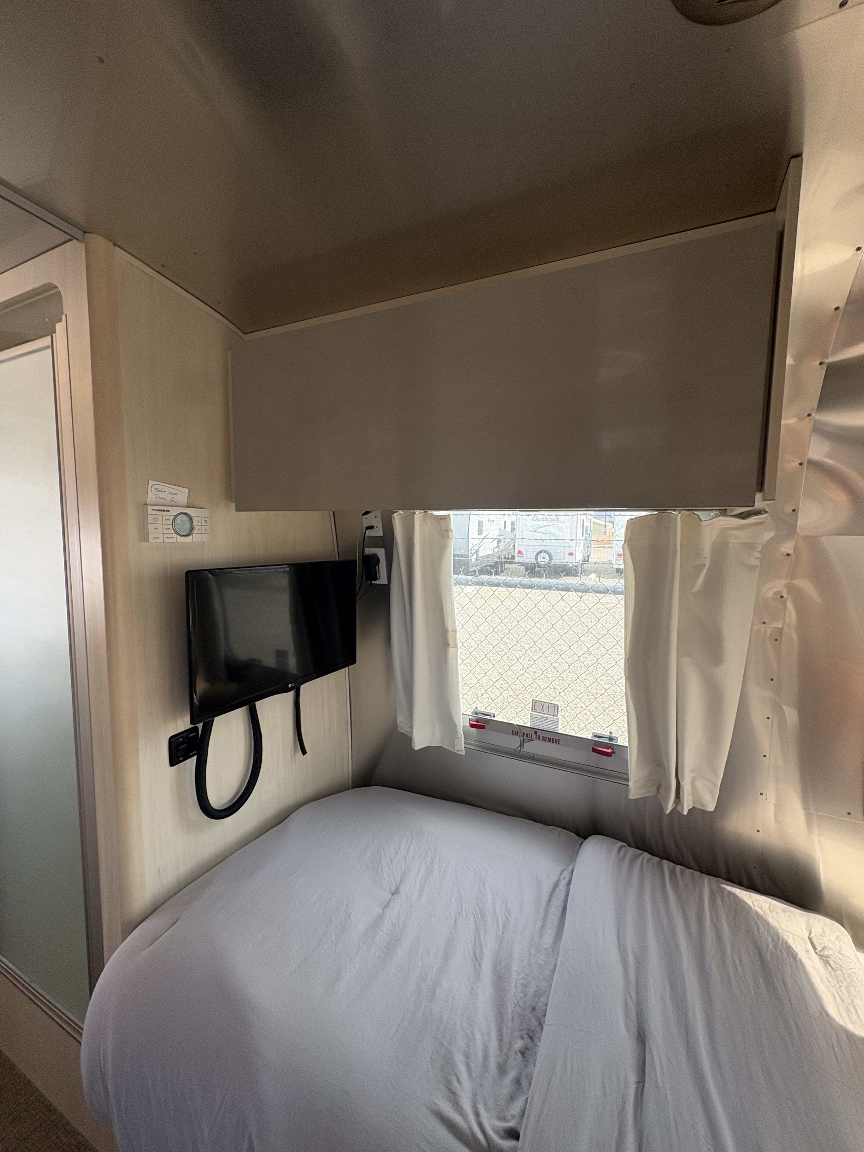 RV rental bedroom: a compact space with a bed, wall-mounted TV, Dometic thermostat, overhead cabinet, and window.. Airstream Flying Cloud 2021