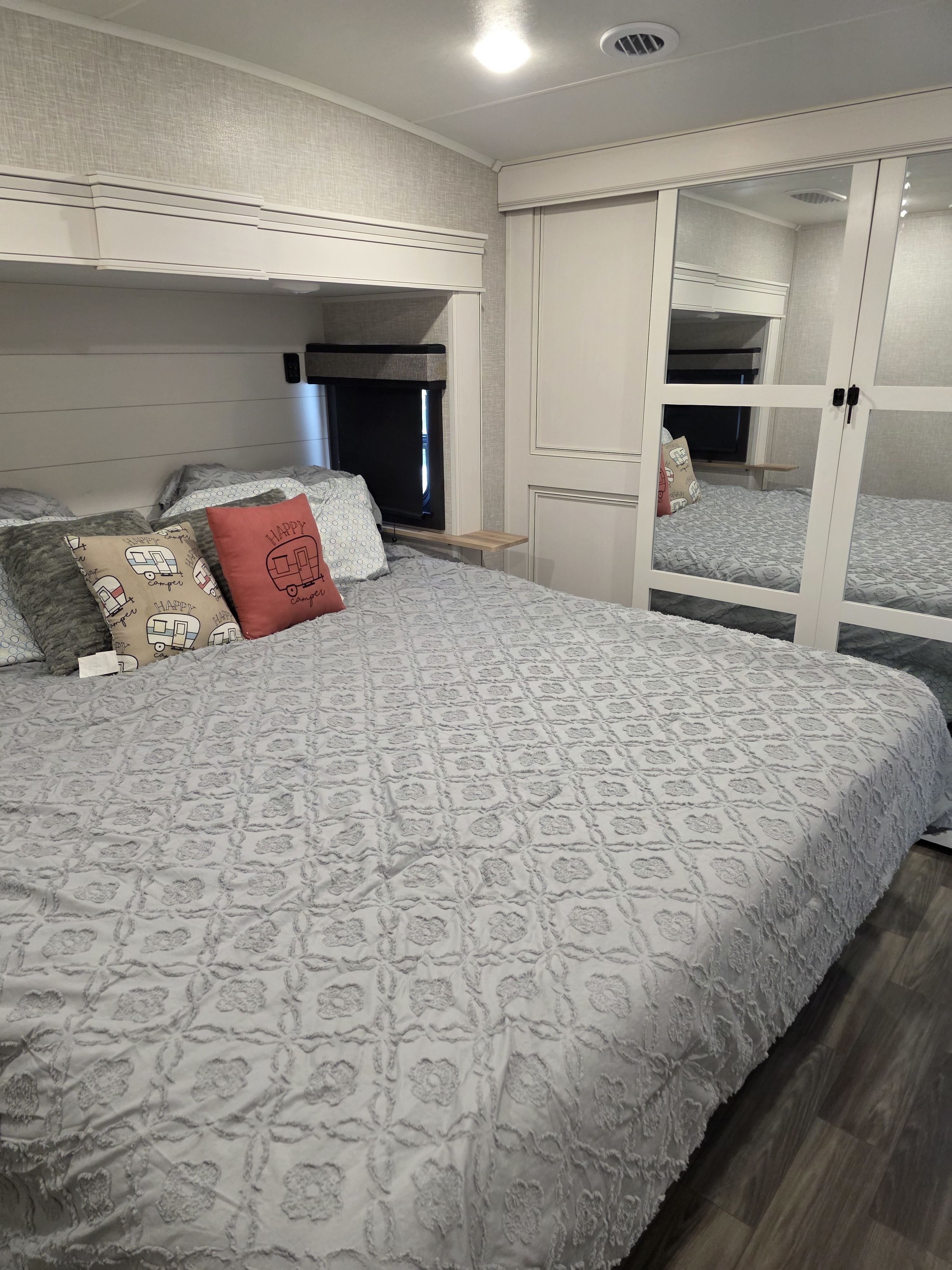 Comfortable RV rental bedroom with a large bed, soft grey comforter, and "Happy Camper" pillows. Mirrored closet and overhead storage.. Jayco Eagle 2024