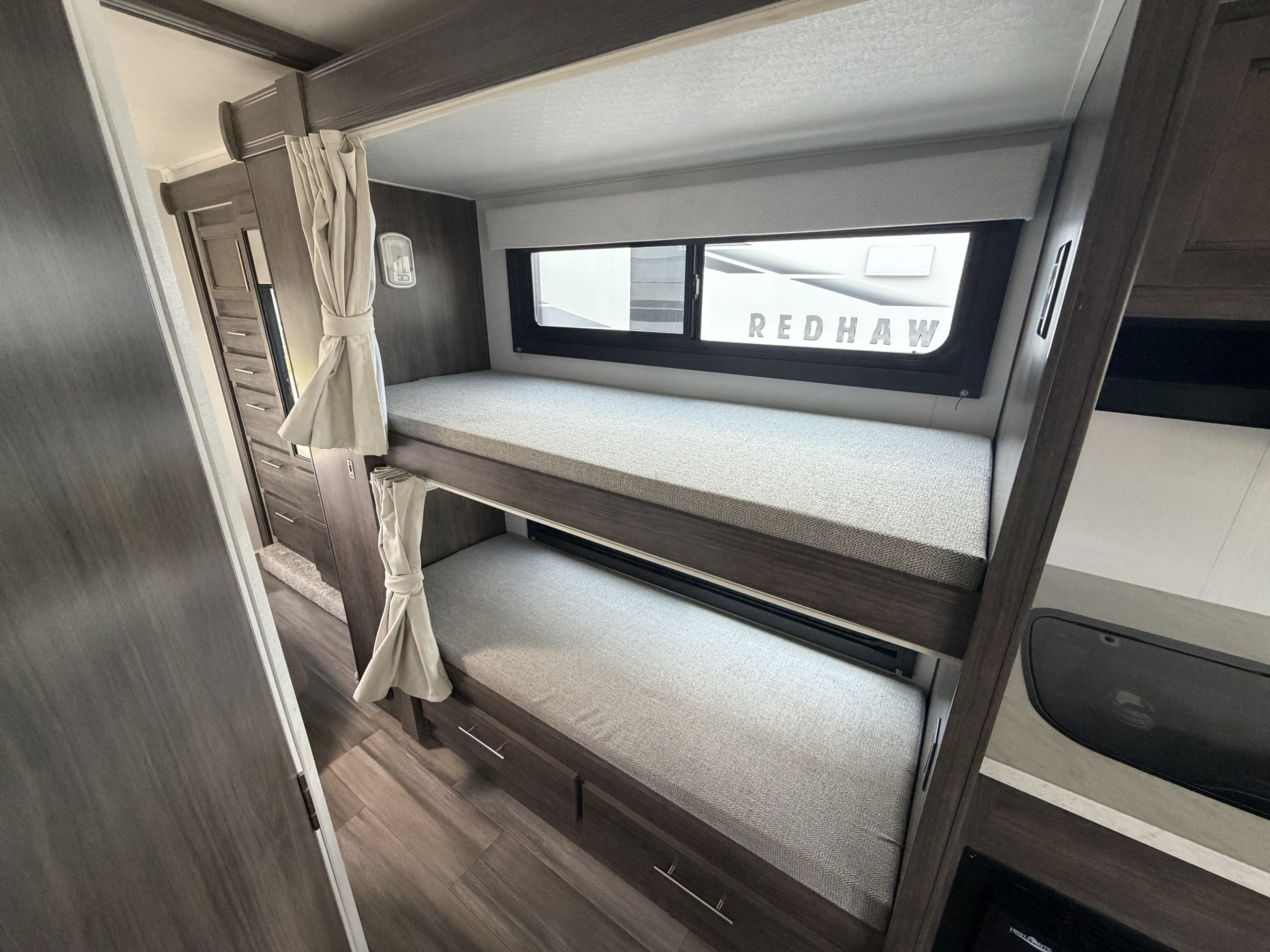 Interior view of an RV rental featuring modern bunk beds with grey cushions and tied-back curtains. A window shows 'REDHAW' branding.. Jayco Redhawk SE 31FF 2026