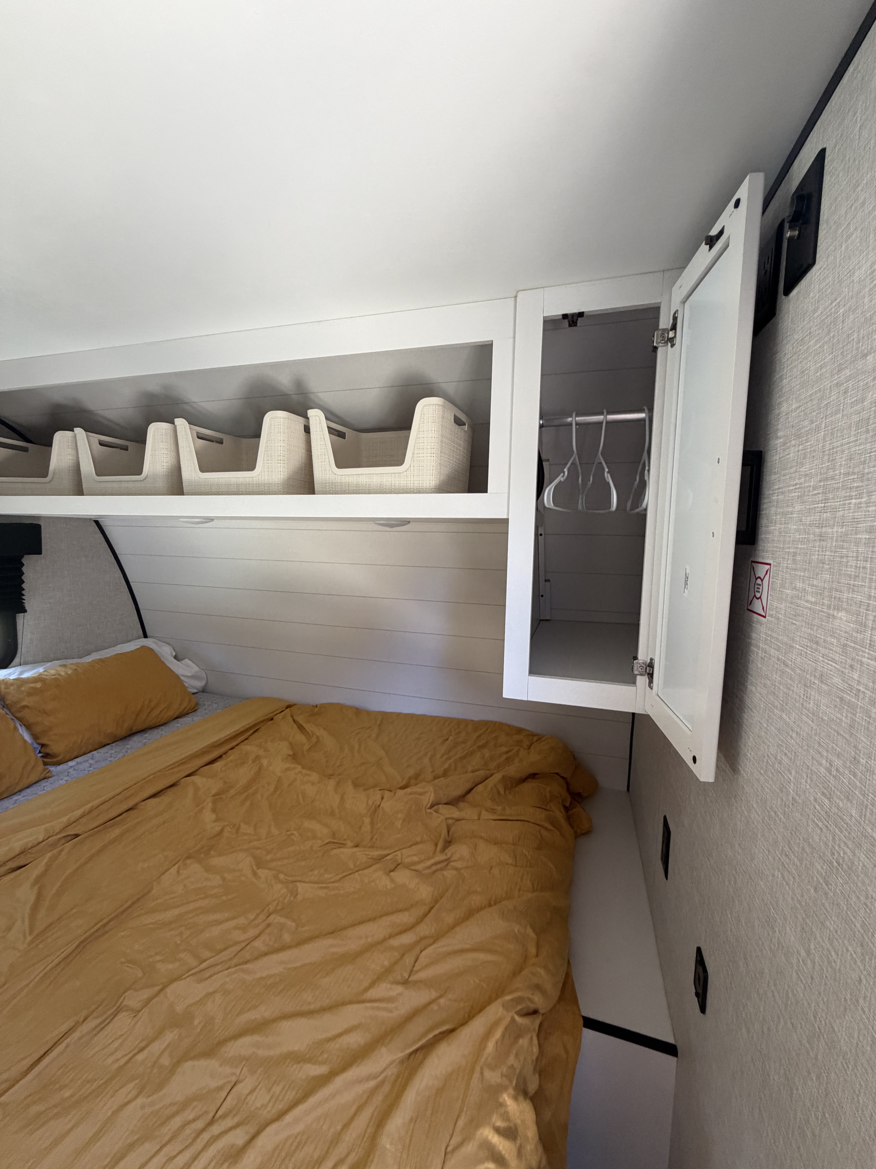 RV rental bedroom: a bed with mustard bedding, overhead storage bins, and an open cabinet with clothes hangers.. Jayco Jay Flight SLX Baja Edition 2025