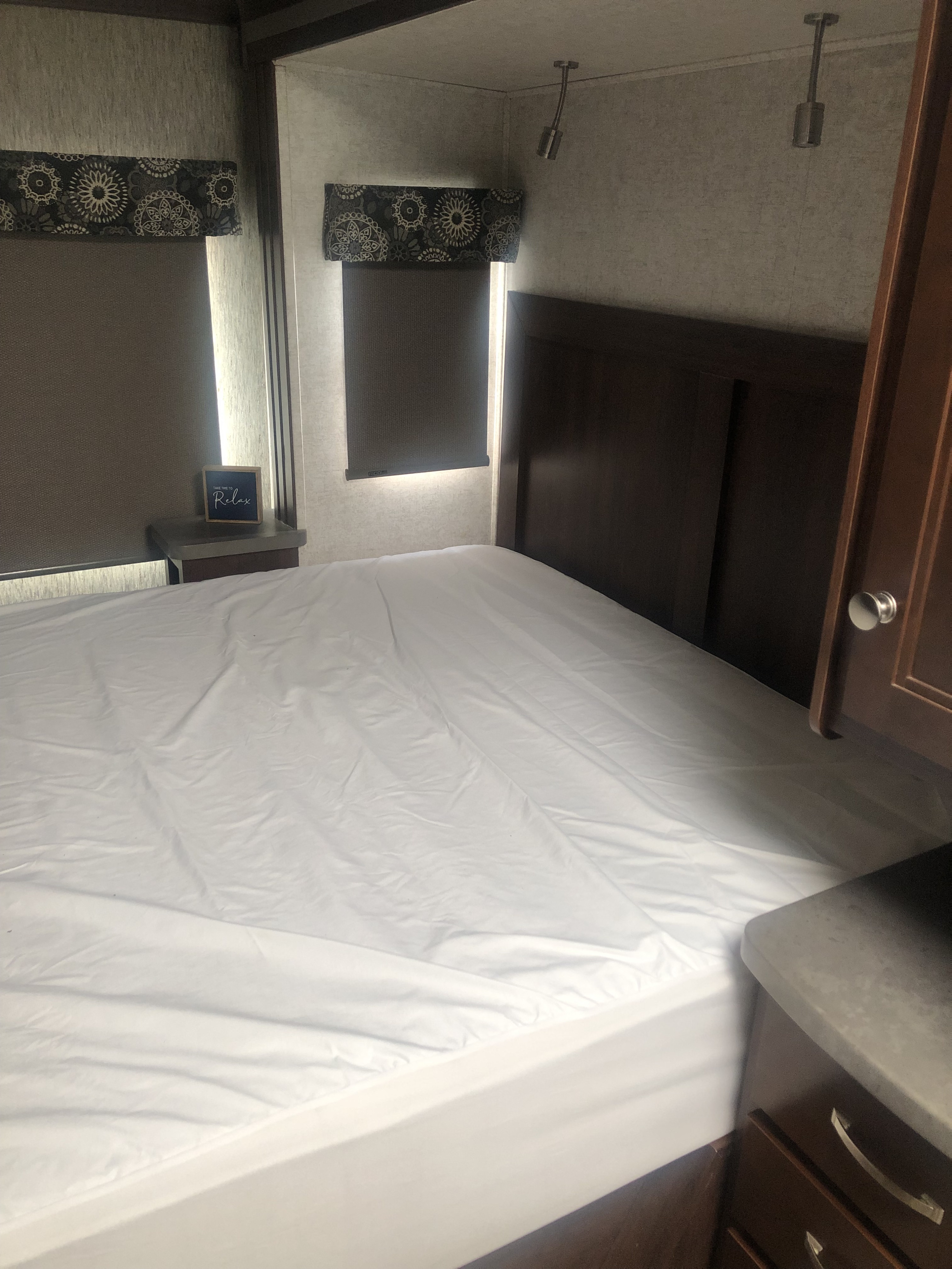 Cozy rv rental bedroom featuring a comfy bed with white sheets, dark wood headboard, and patterned window shades.. Forest River Sunseeker 2017