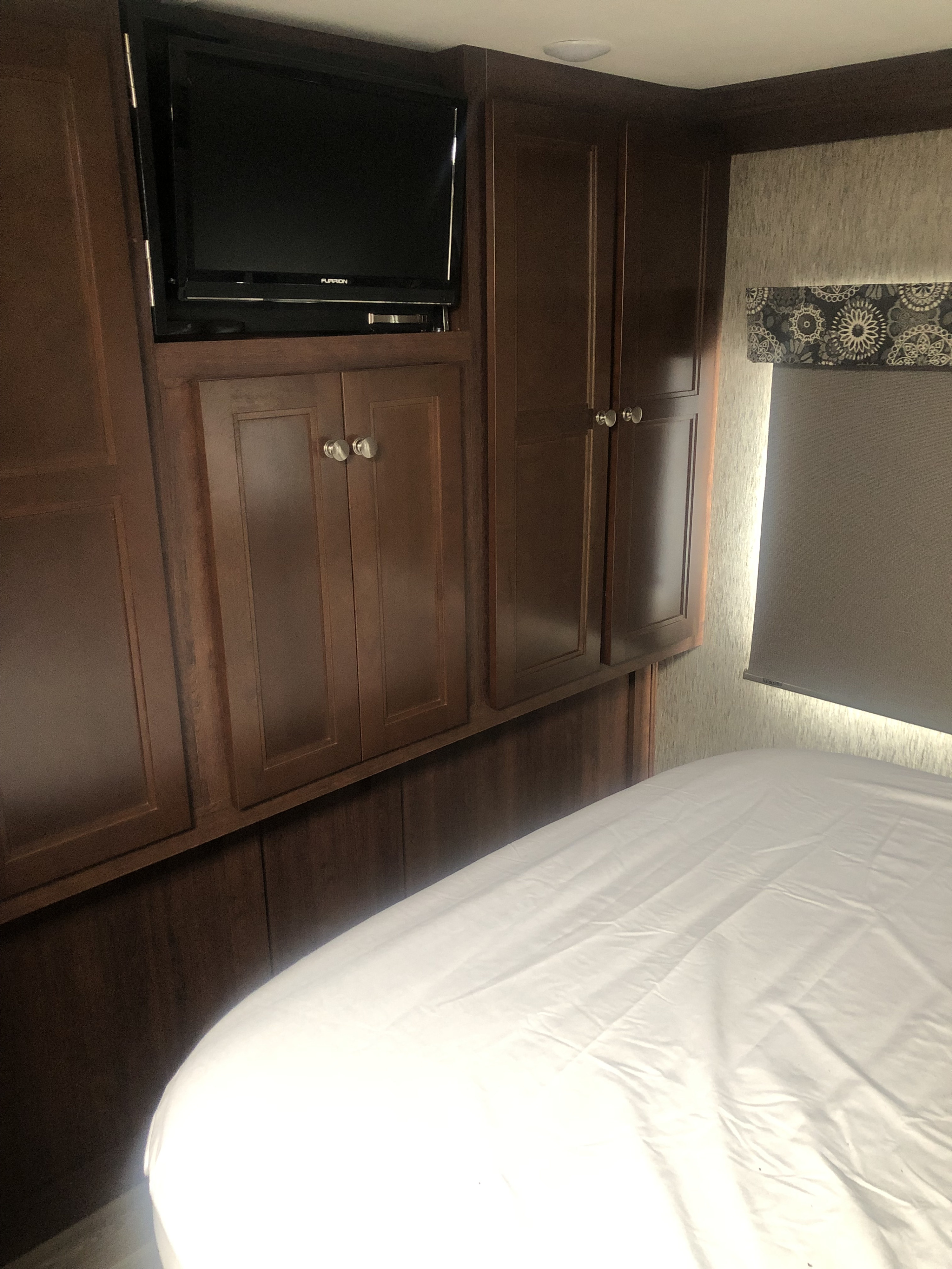 RV rental bedroom featuring a bed with white sheets, dark wood cabinetry, a mounted Furrion TV, and a window with blinds.. Forest River Sunseeker 2017
