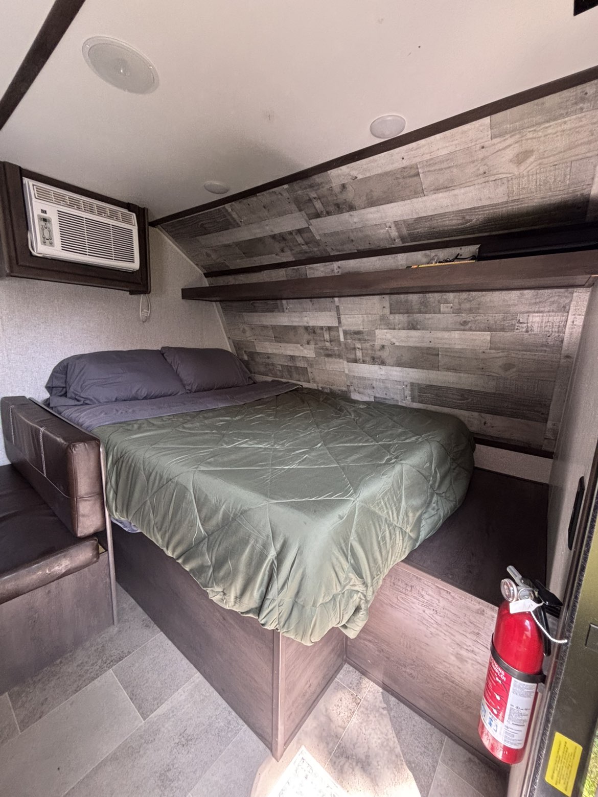 Cozy rv rental bedroom with a bed, AC unit, and shelves. A fire extinguisher is mounted near the door, featuring wood-look walls.. Dutchmen Coleman Lantern LT 2021