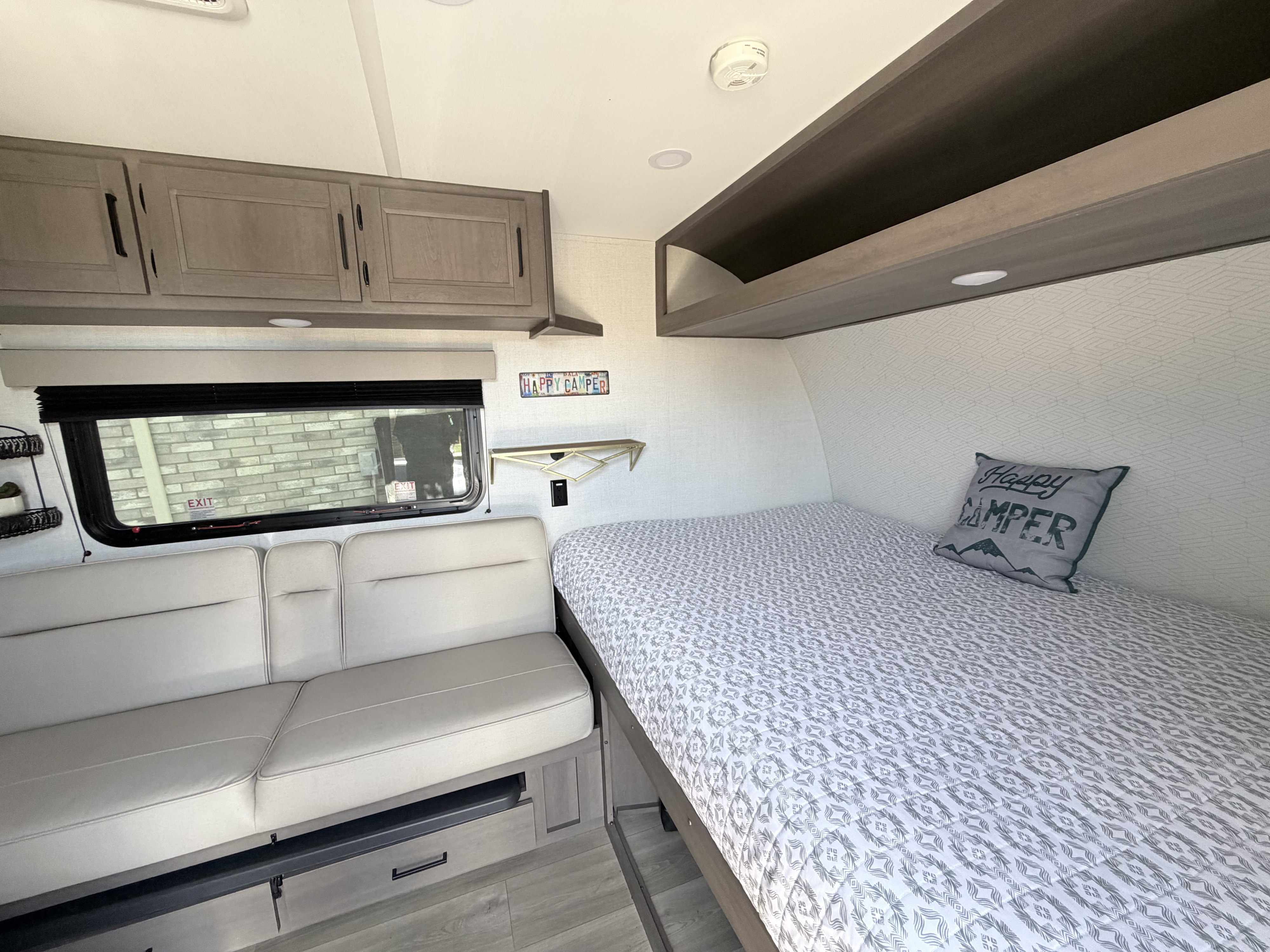 Cozy RV rental interior with a patterned bed, "Happy Camper" pillow, light sofa, and overhead cabinets for compact living.. Grand Design Transcend One 2025