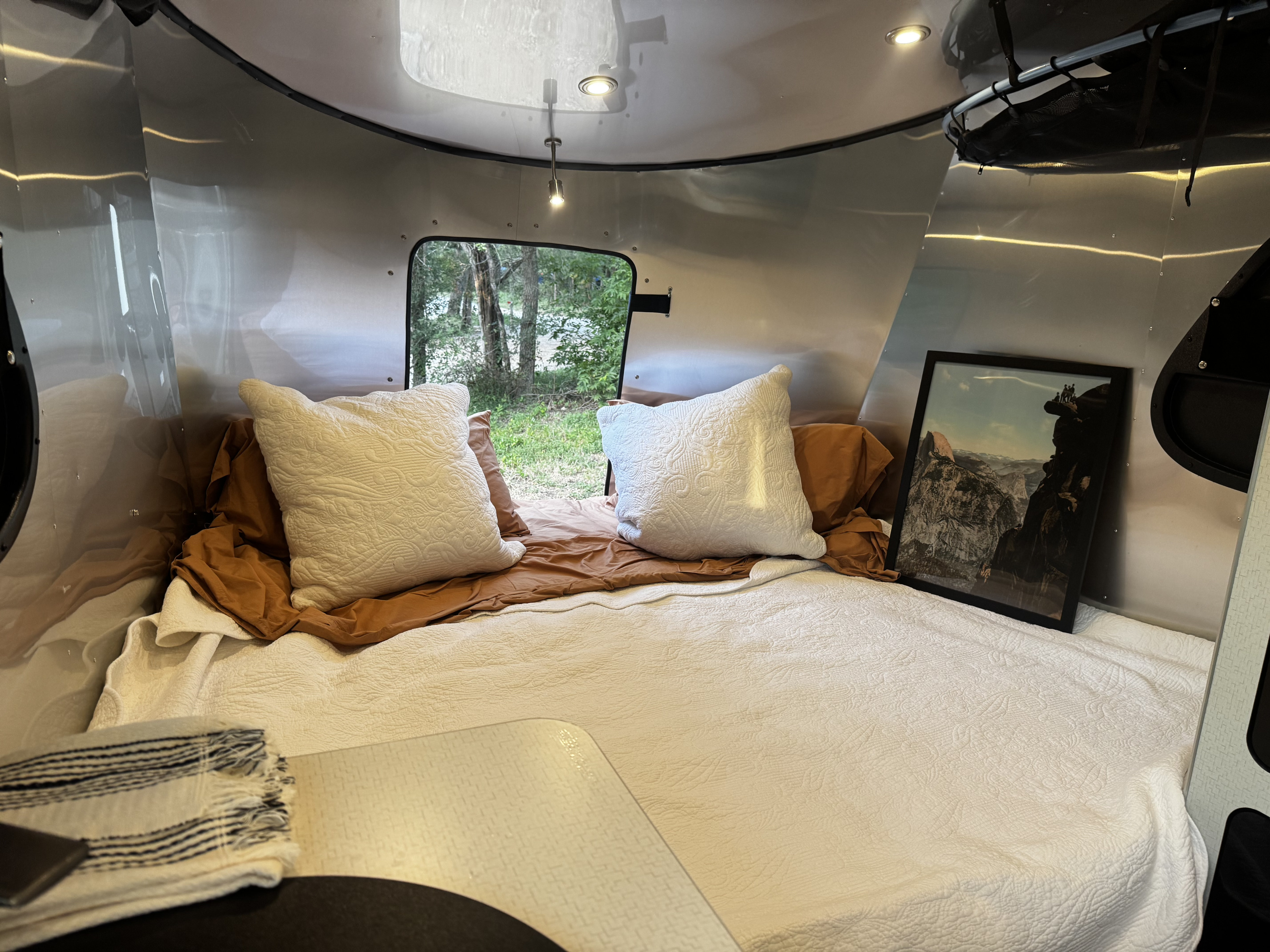 Cozy RV rental interior with a comfy bed, white bedding, decorative pillows, and a scenic window view of nature.. Airstream Basecamp 2024