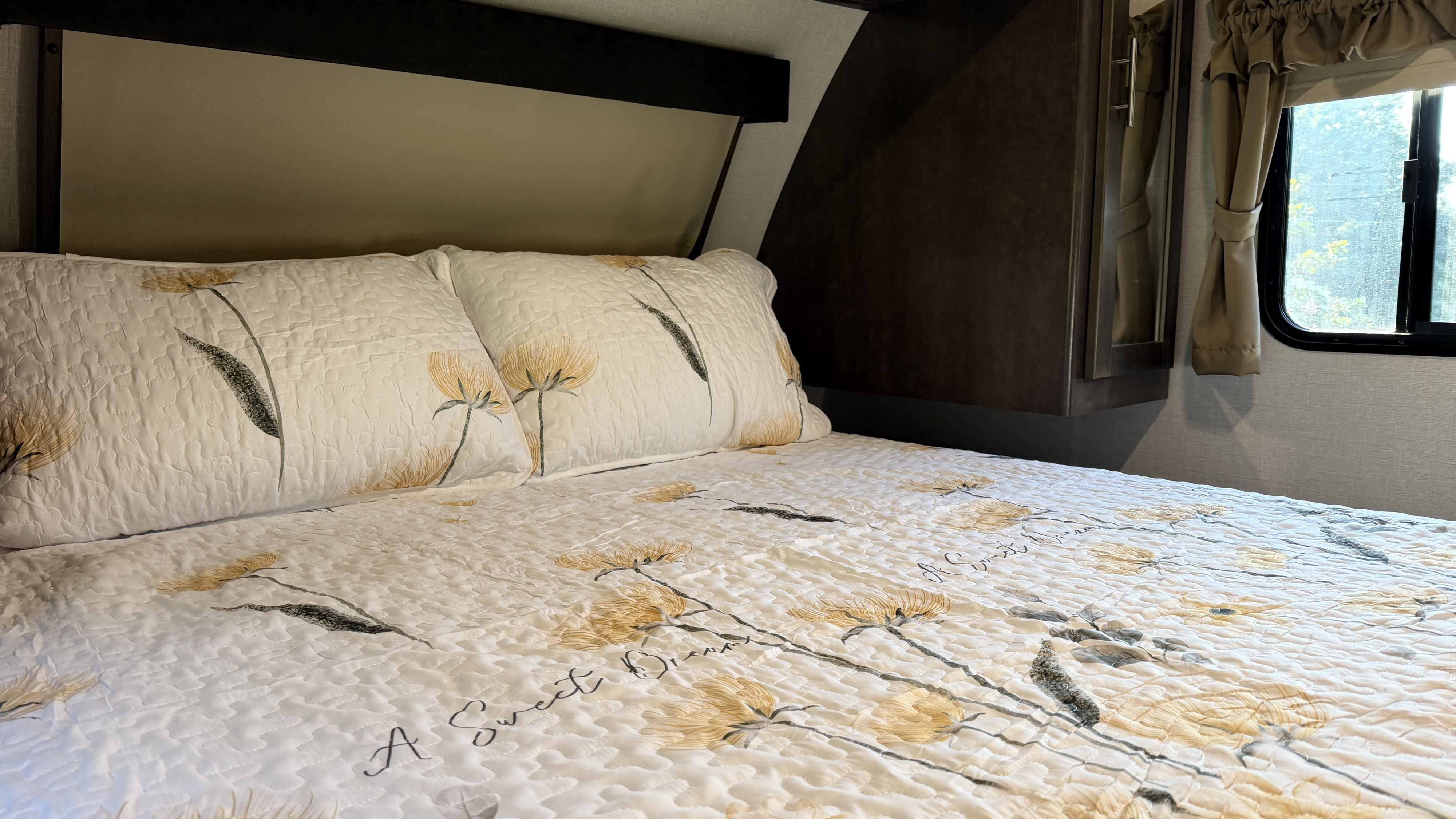 Cozy bed with floral quilt and "A Sweet Dream" message in an RV rental. Dark wood cabinetry & window with curtains visible.. Crossroads RV Sunset Trail Grand Reserve 2017