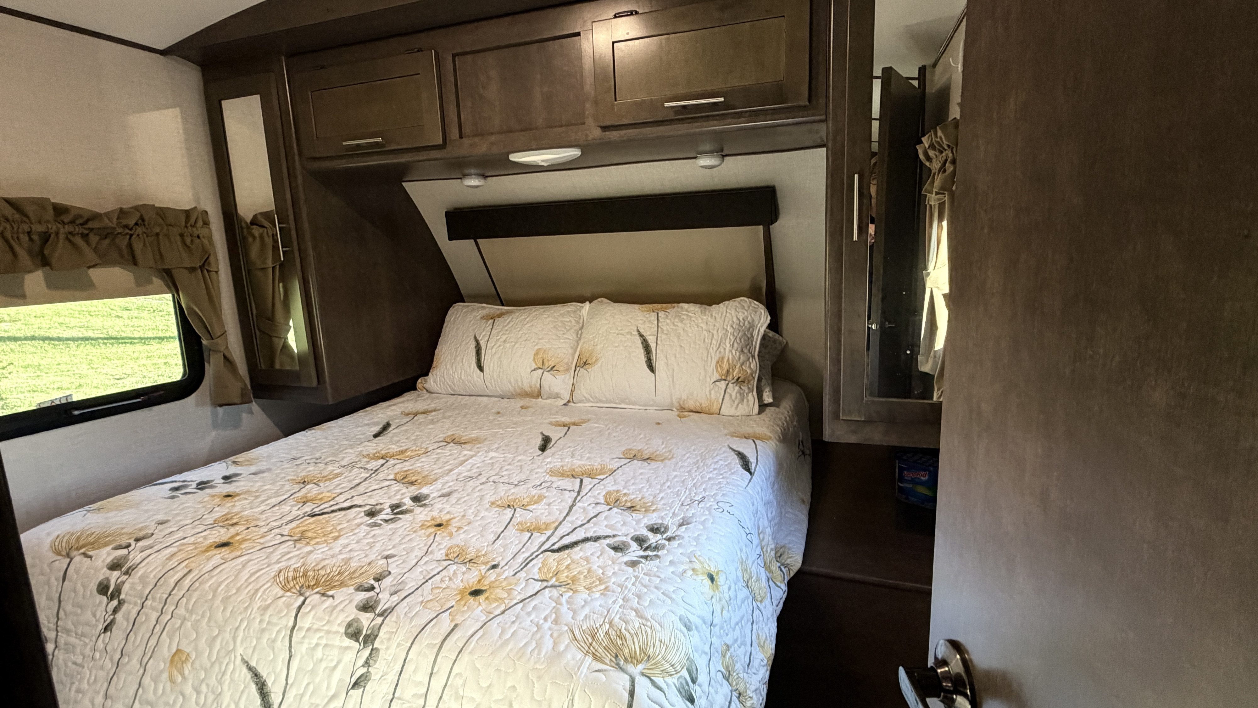 Cozy RV rental bedroom with a comfy bed, floral bedding, overhead storage, and a window view, ideal for travel.. Crossroads RV Sunset Trail Grand Reserve 2017