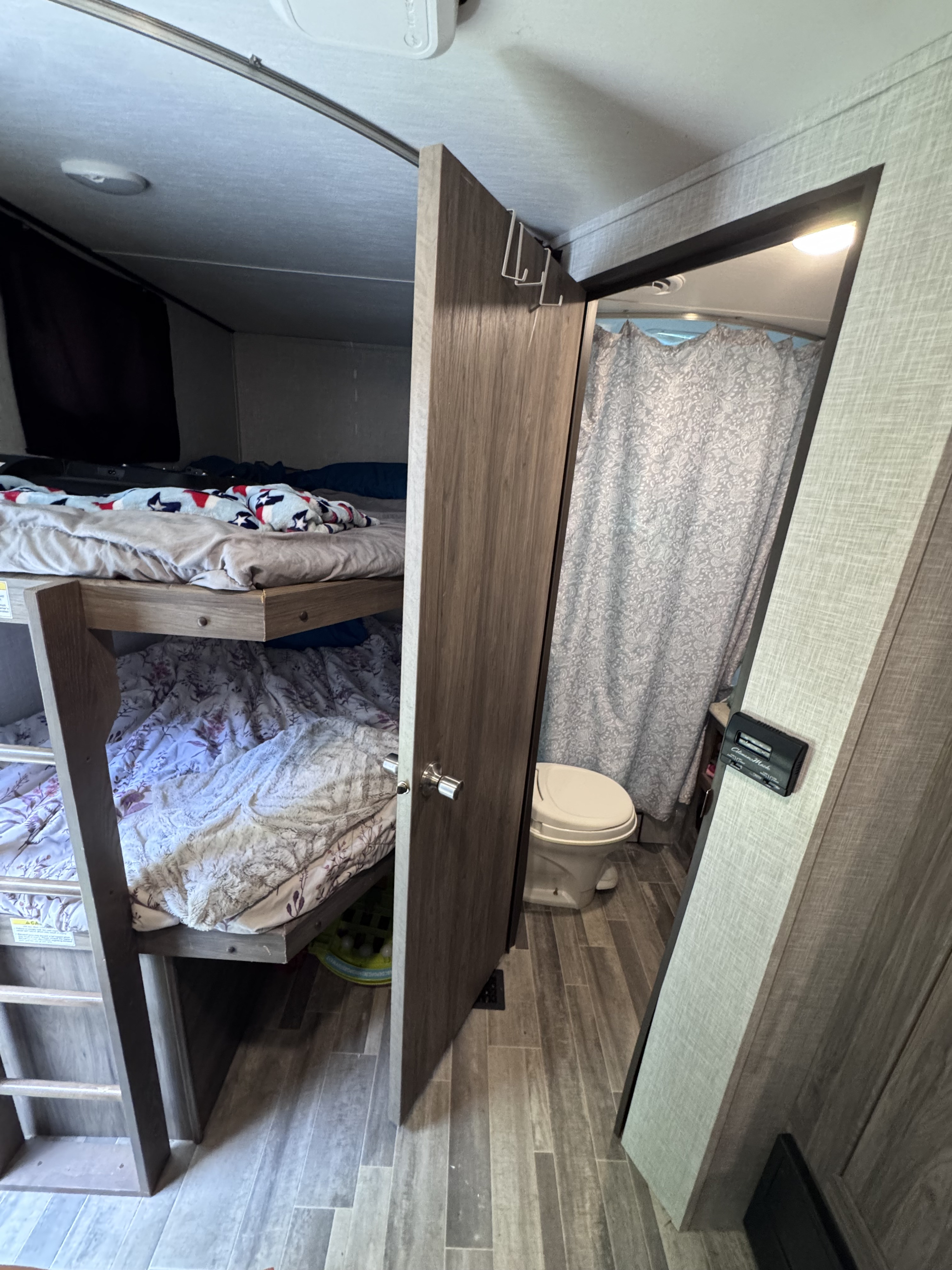 RV rental interior view: bunk beds with soft blankets on the left, a wooden door opens to a bathroom with a toilet and shower.. Crossroads RV Zinger Lite 2022