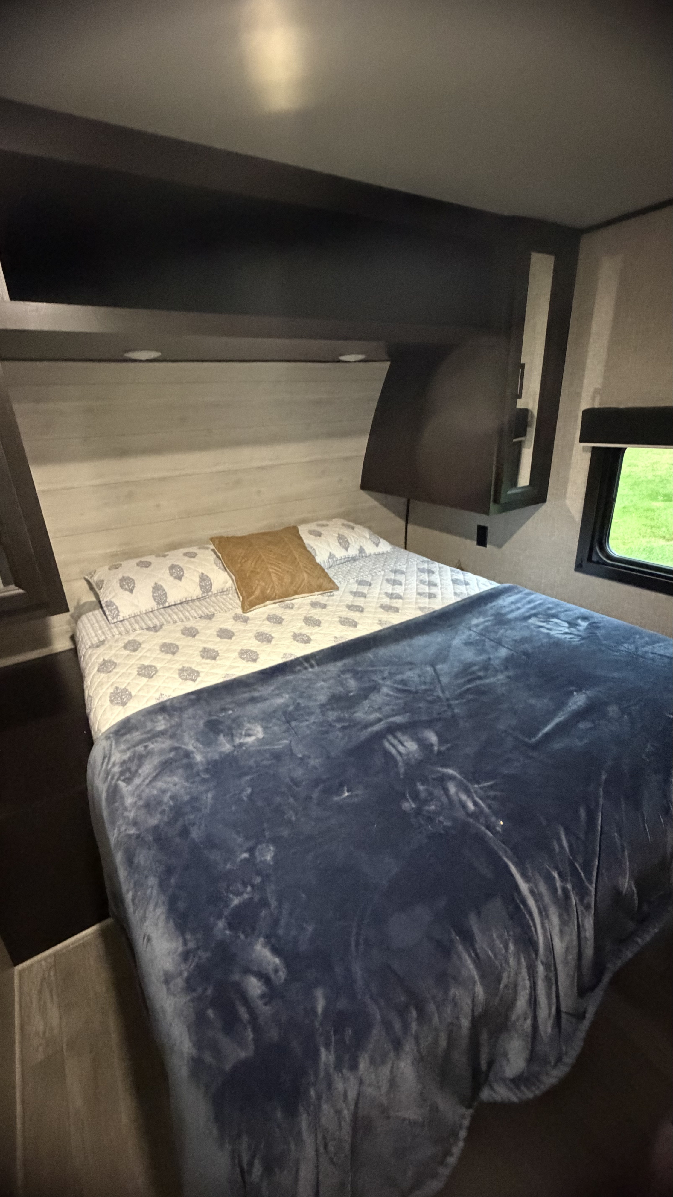 Cozy RV rental bedroom with a comfy bed, blue blanket, light wood headboard, overhead cabinets, and a window.. Jayco Jay Flight 2023