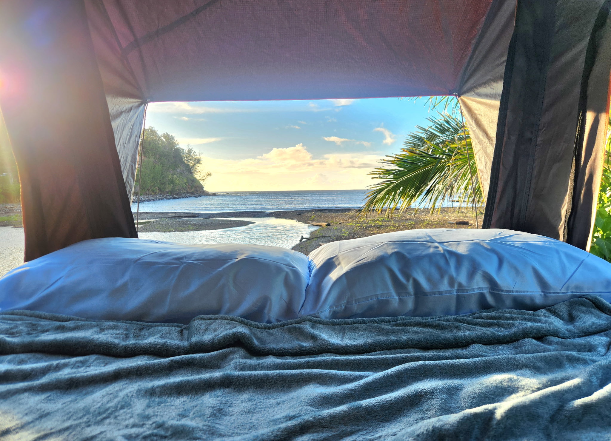 Cozy RV rental bed with white pillows and a grey blanket, overlooking a serene beach, ocean, green headland, and palm tree.. Subaru Outback 2017