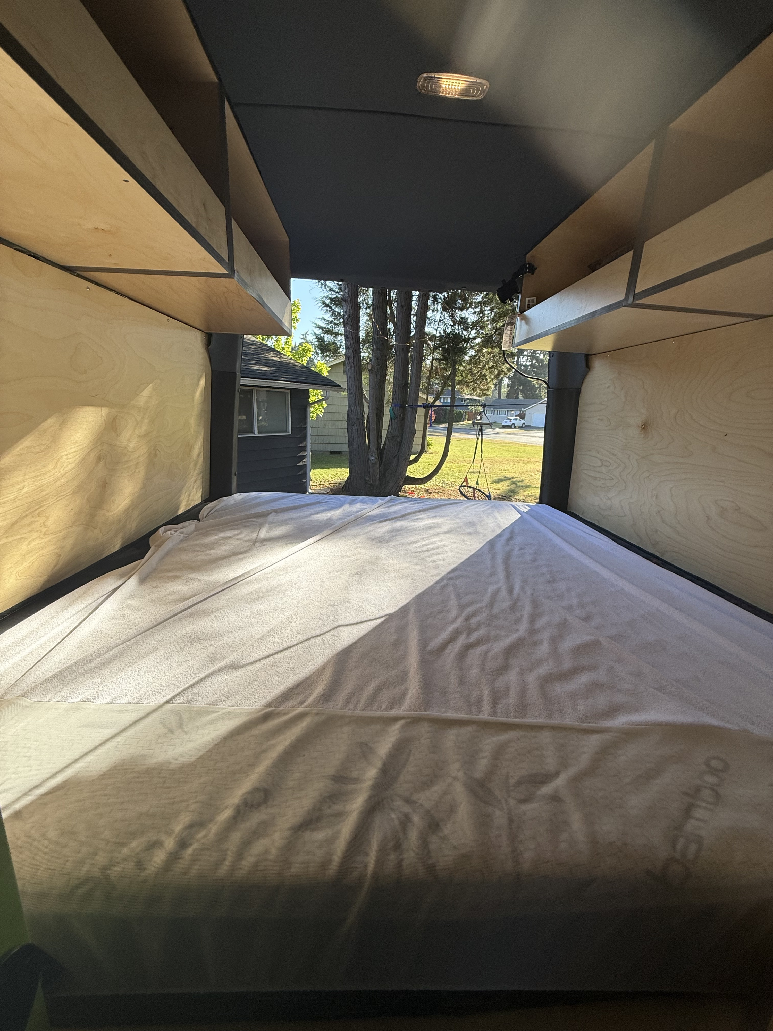 RV rental interior: a comfortable bed with white sheets, wooden cabinets, and a clear view of trees and a house outside.. Mercedes-Benz Sprinter 2024