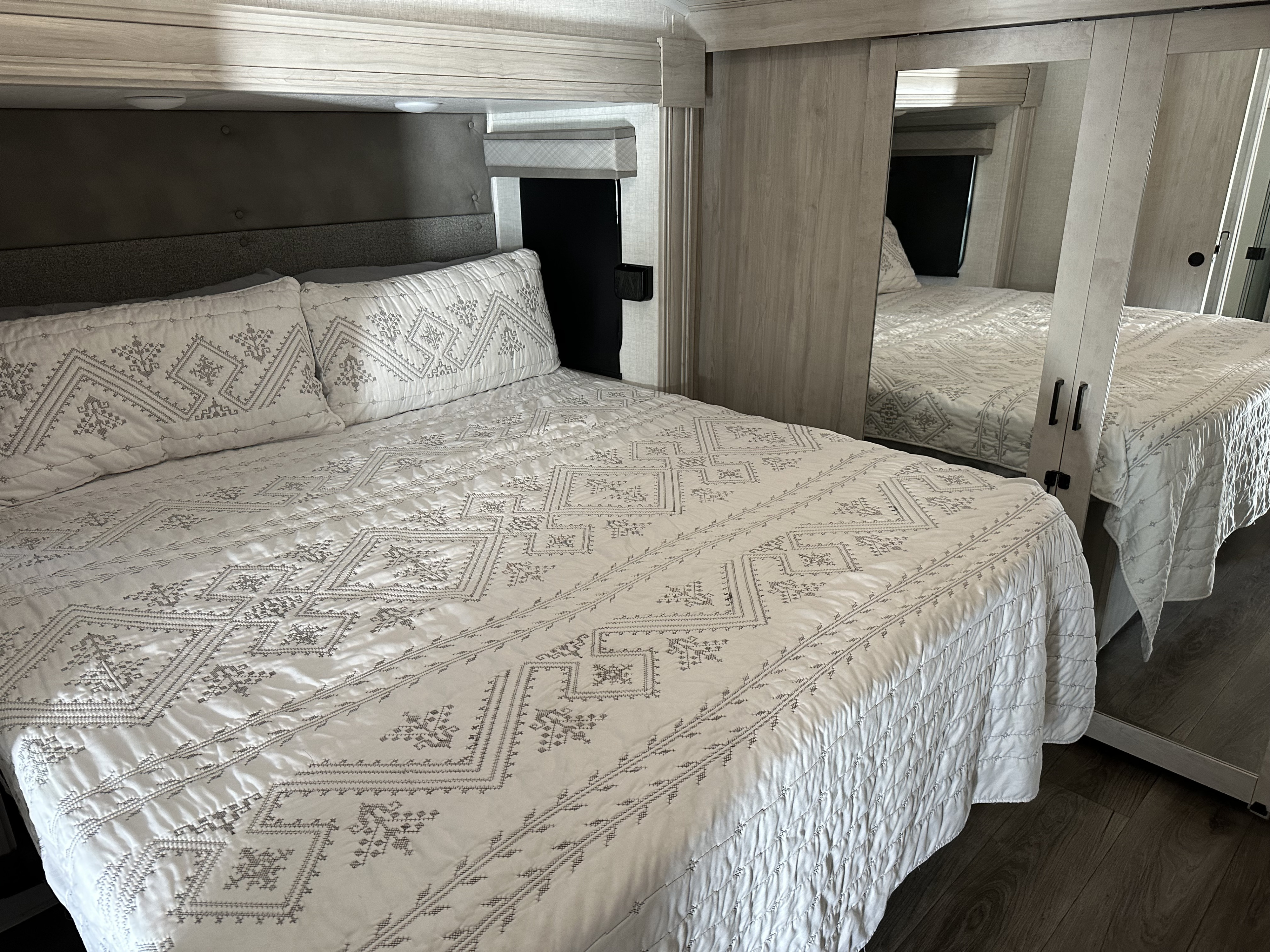 Cozy RV rental bedroom: bed with white/gray patterned quilt, upholstered headboard, light wood trim, mirrored closet.. East to West Tandara 2022