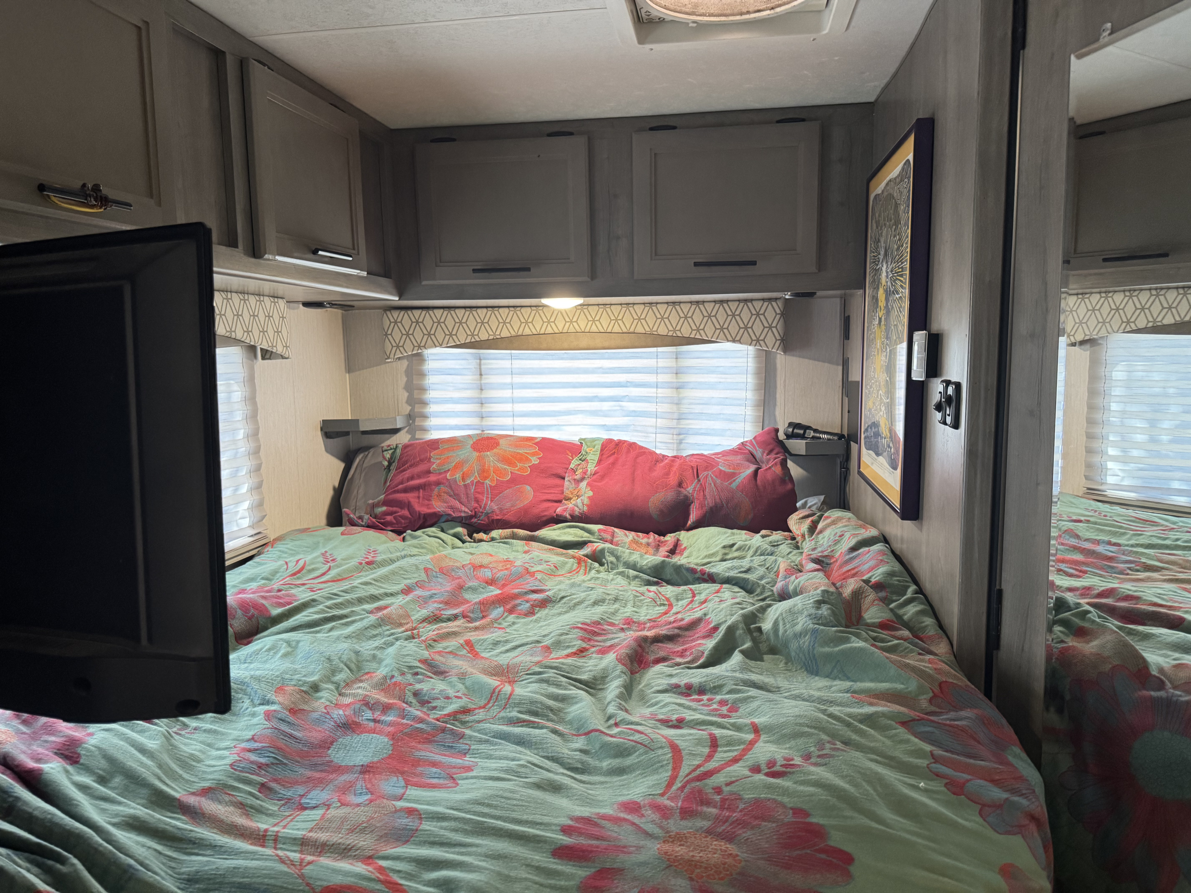 Cozy RV rental bedroom with vibrant floral bedding, overhead cabinets, window blinds, and a TV, providing a comfortable space.. Forest River Coachmen Freelander 2021