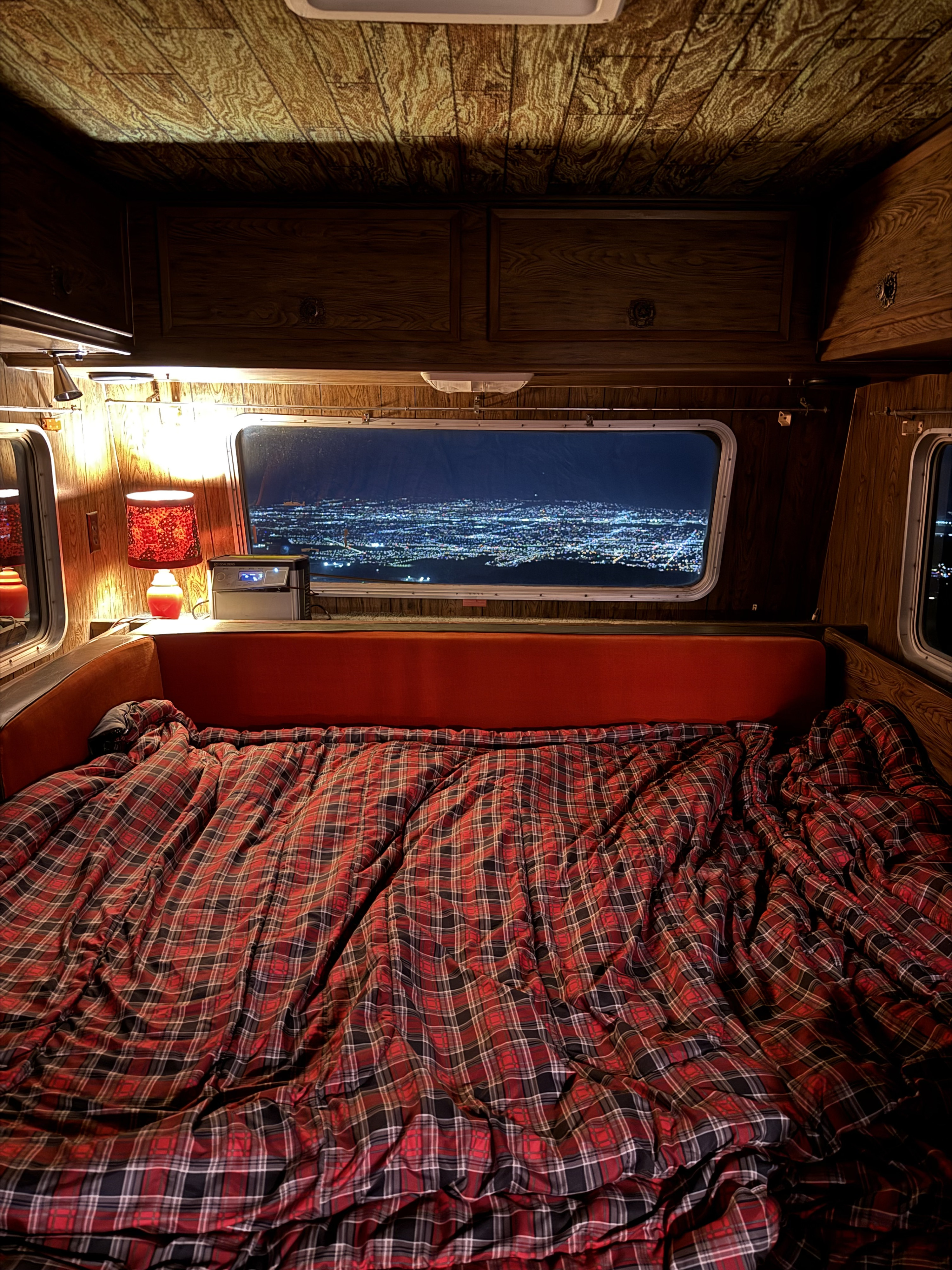 Cozy RV rental interior at night, featuring a red plaid bed, warm lamp, and a stunning city lights view from the window.. International 510A 1973