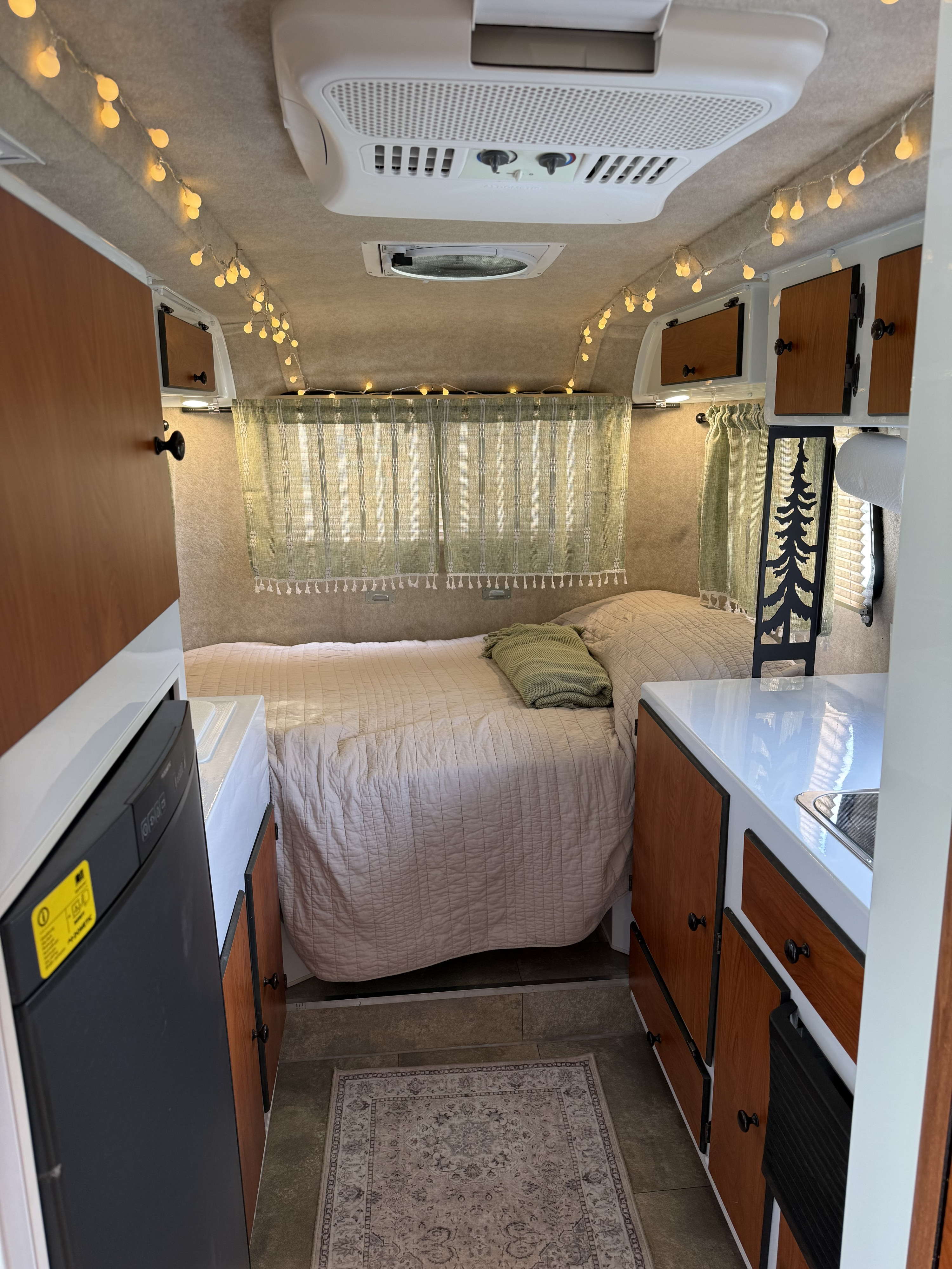 Inside a charming RV rental: full bed with quilt, string lights, wood cabinets, sink, fridge, and green curtains.. Scamp Scamp Trailer 2022