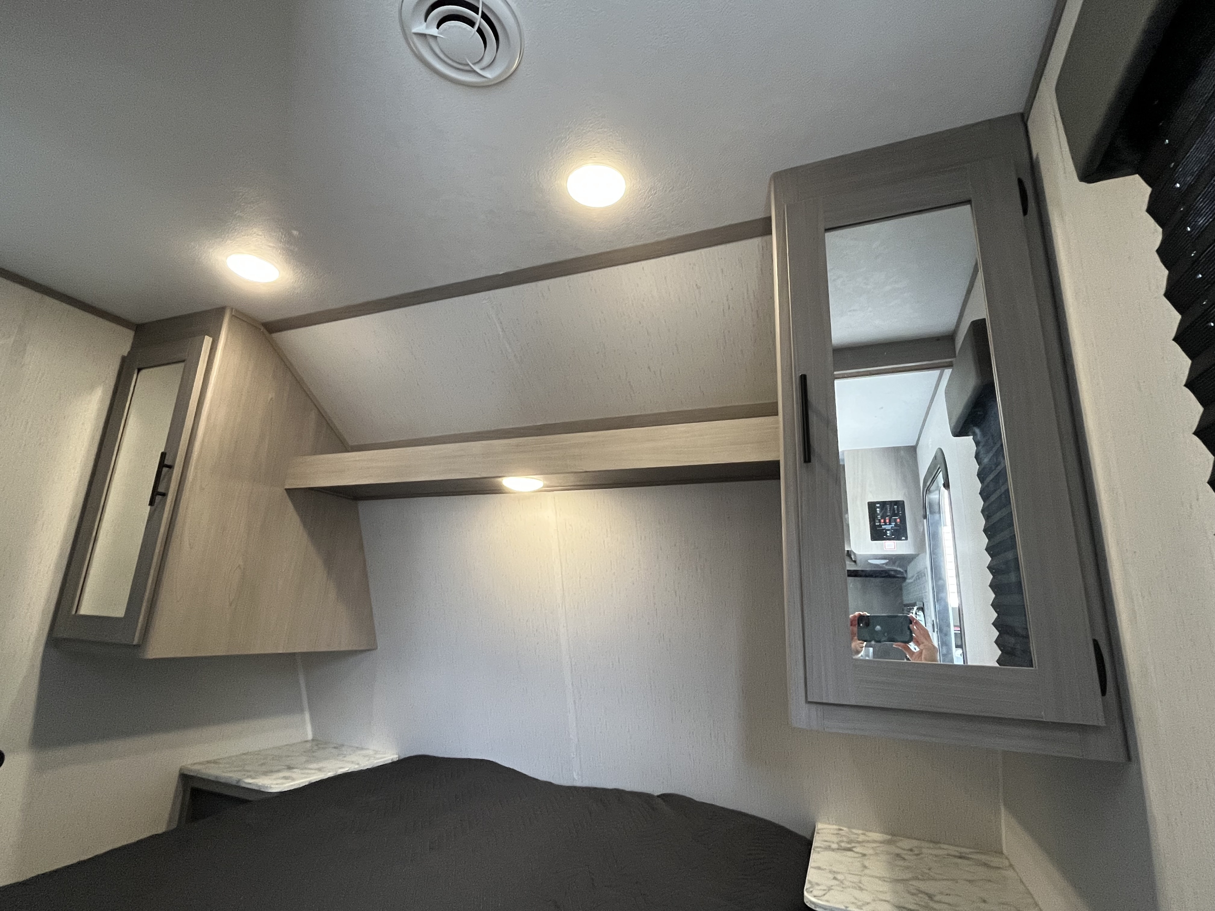 Inside an RV rental, a bedroom features overhead lighting, storage cabinets, and a comfortable bed. Photographer visible in mirror.. Dutchmen Coleman Lantern LT 2022