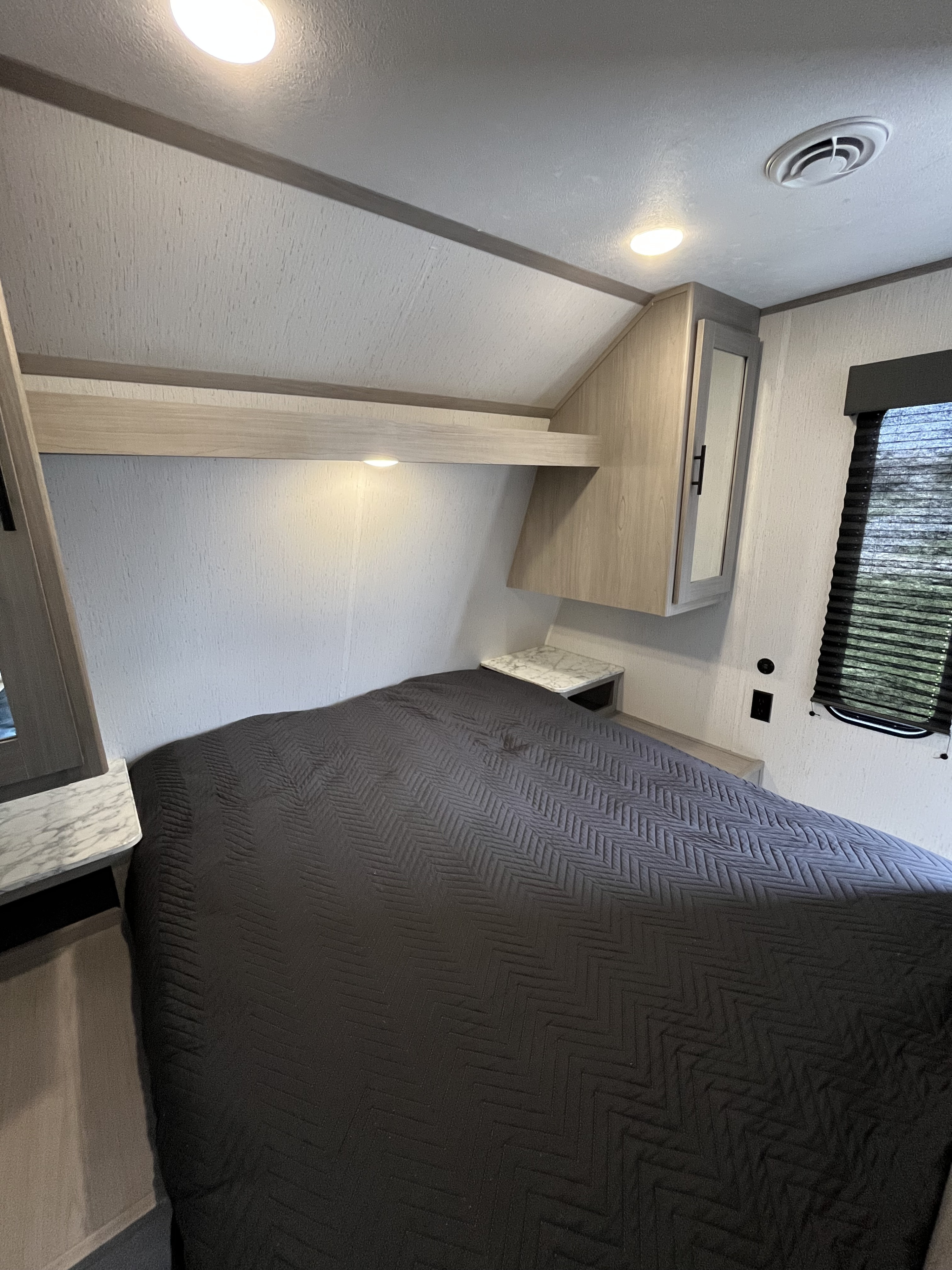 RV rental bedroom: a modern space with a dark gray quilted bed, overhead cabinets, small side tables, and a window with blinds.. Dutchmen Coleman Lantern LT 2022