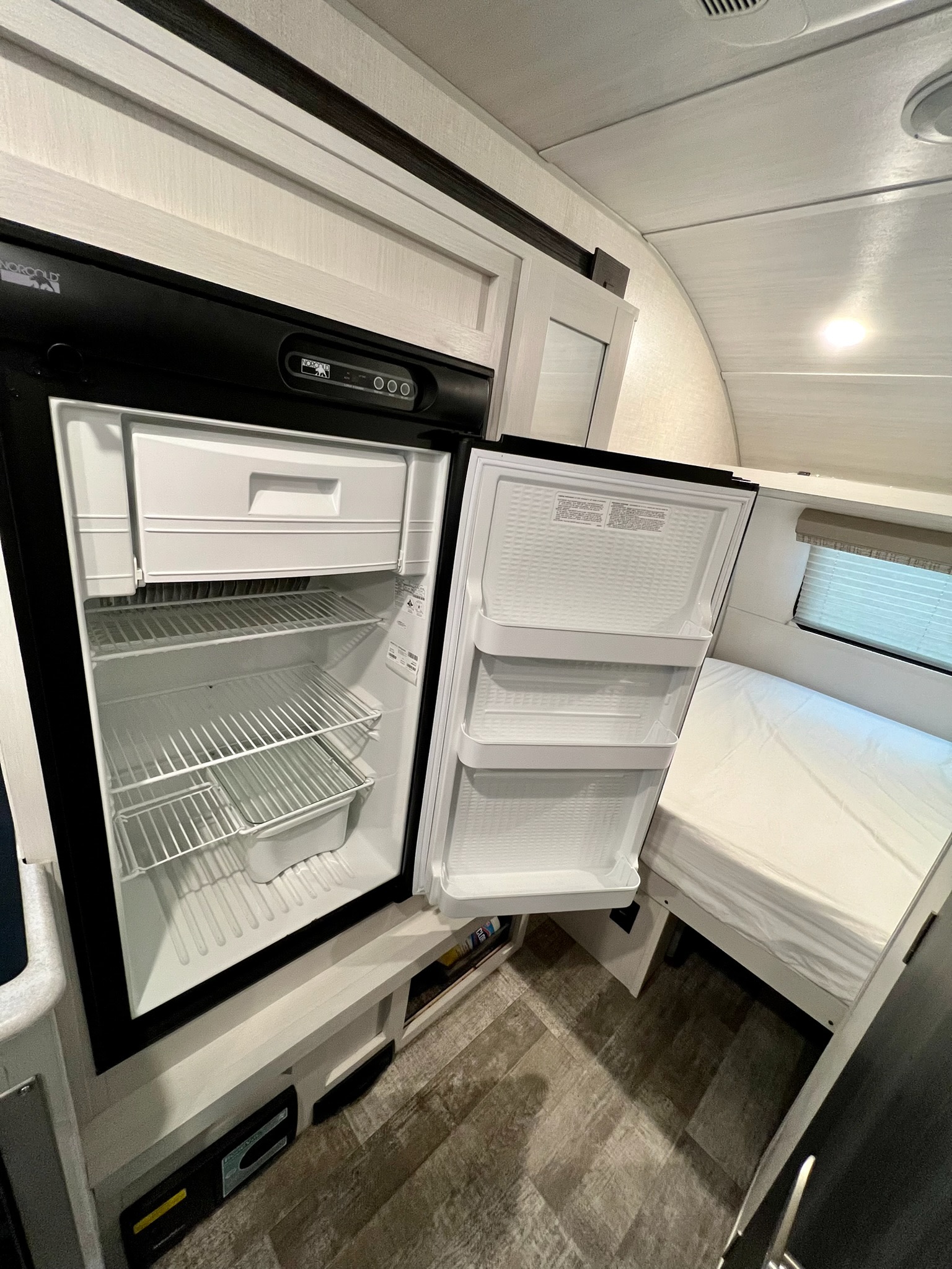 Open Norcold refrigerator and freezer in an RV rental, revealing compact design next to a bed and window.. Forest River R-Pod 190 2021