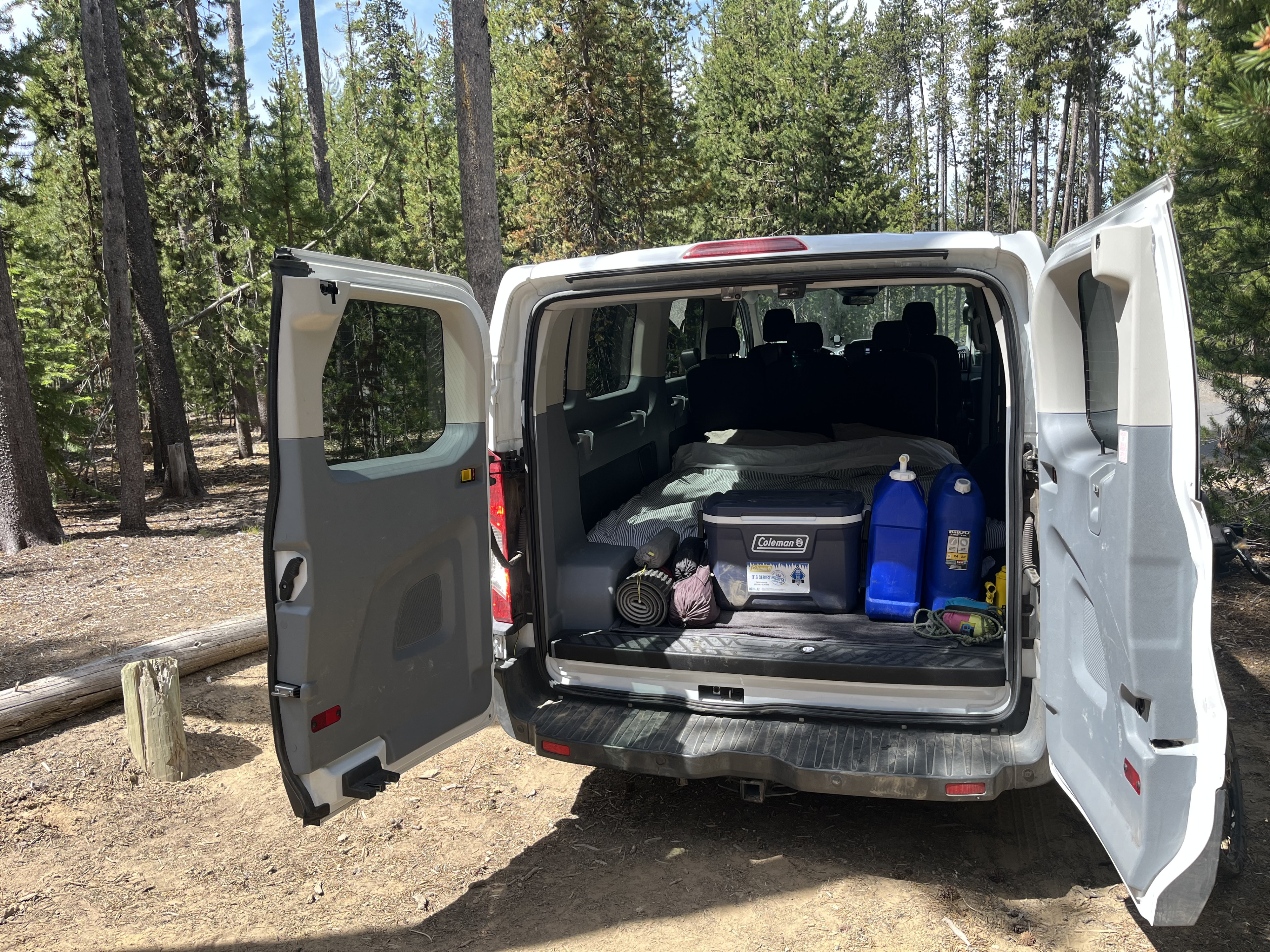 RV rental van packed for camping in the forest, featuring a bed, cooler, and water jugs. Ready for adventure!. ford transit 2017