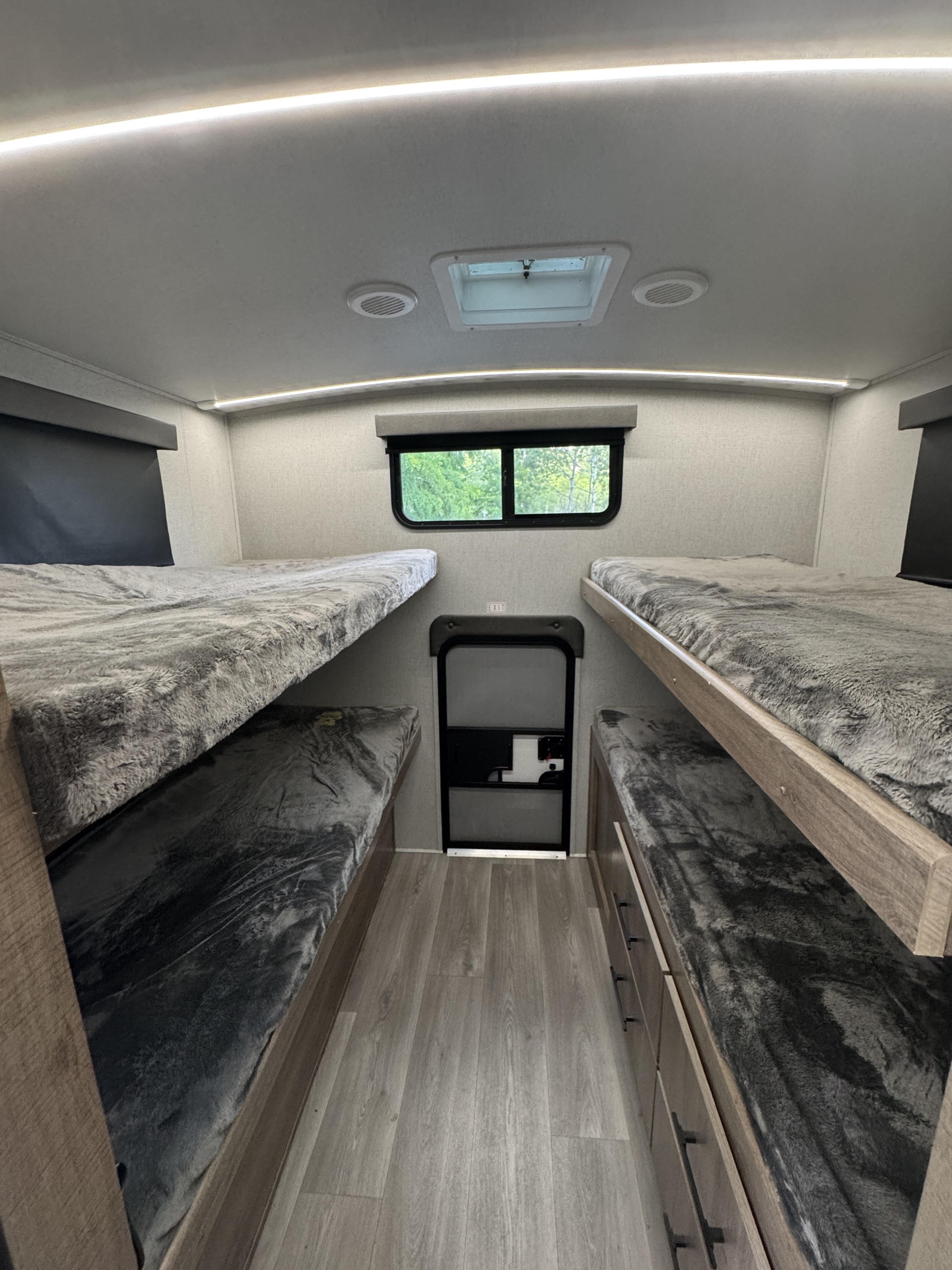 Inside an RV rental: two sets of bunk beds with fluffy gray bedding, light wood floor, rear window, and emergency exit.. Grand Design Imagine 2024