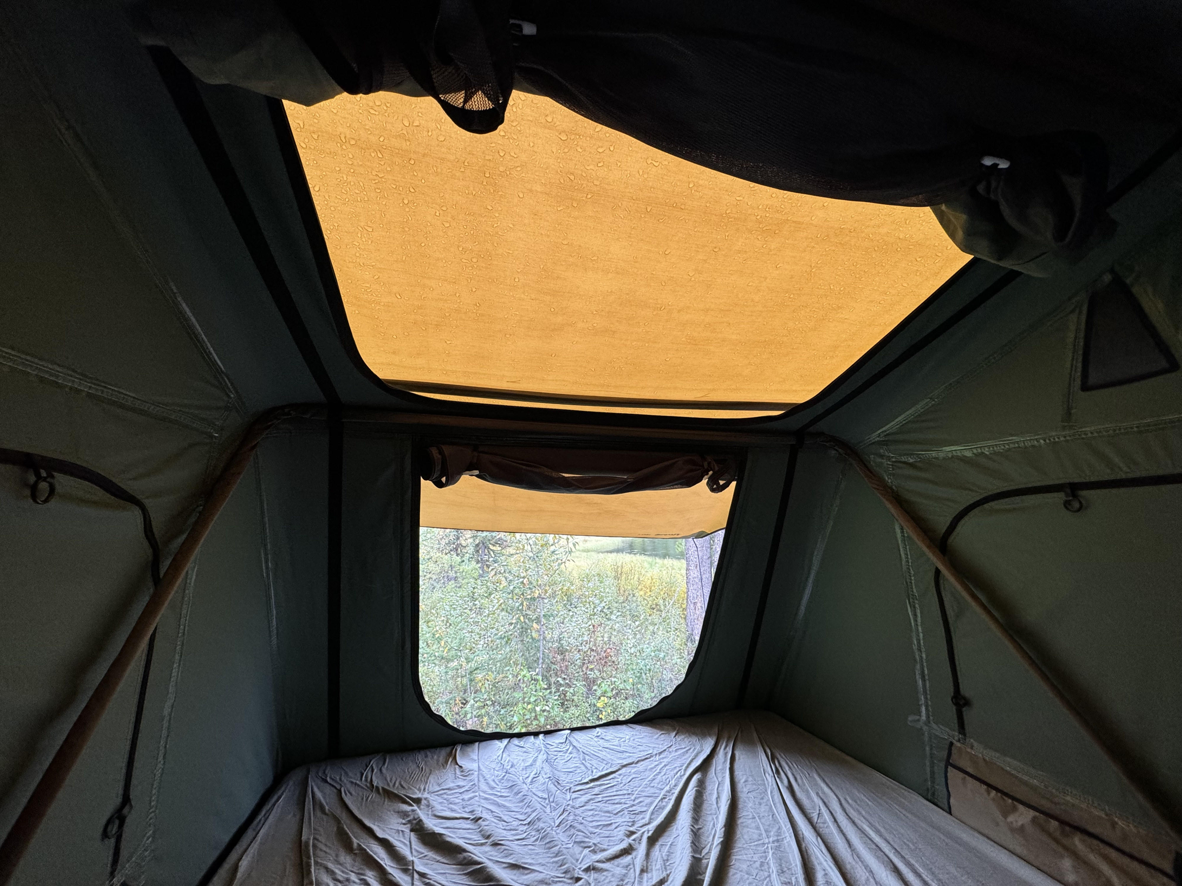 Inside an rv rental rooftop tent. Raindrops visible on the roof, a cozy bed, and a green nature view through the window.. Volvo XC60 2022