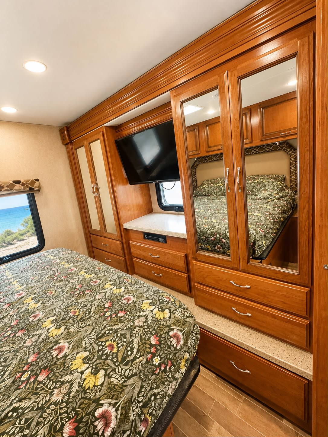 Cozy RV rental bedroom featuring floral bedding, wooden cabinetry, mounted TV, and mirrored closet doors. A window overlooks an ocean view.. Jamboree Jamboree Motorhome 2018