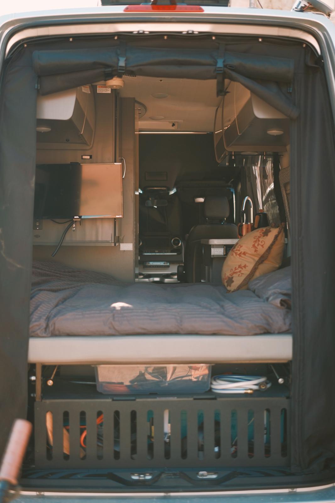 Cozy RV rental interior, viewed from the back, showing bed, TV, kitchen sink, and organized storage. A perfect campervan setup.. Thor Tranquility 2023