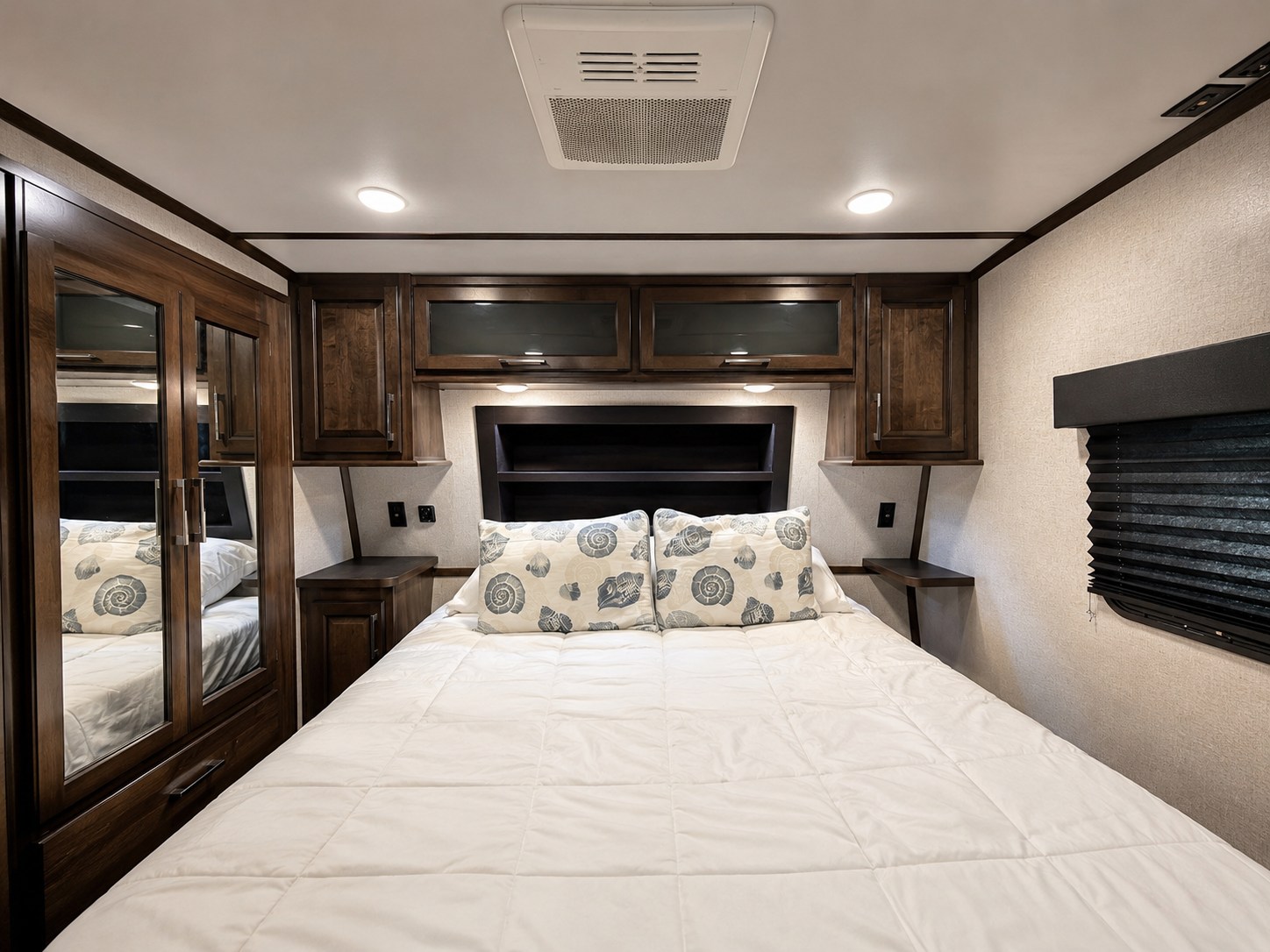 A modern RV rental bedroom featuring a queen bed with seashell pillows, dark wood storage, and bright lighting.. Dutchmen Triton 4271 2022