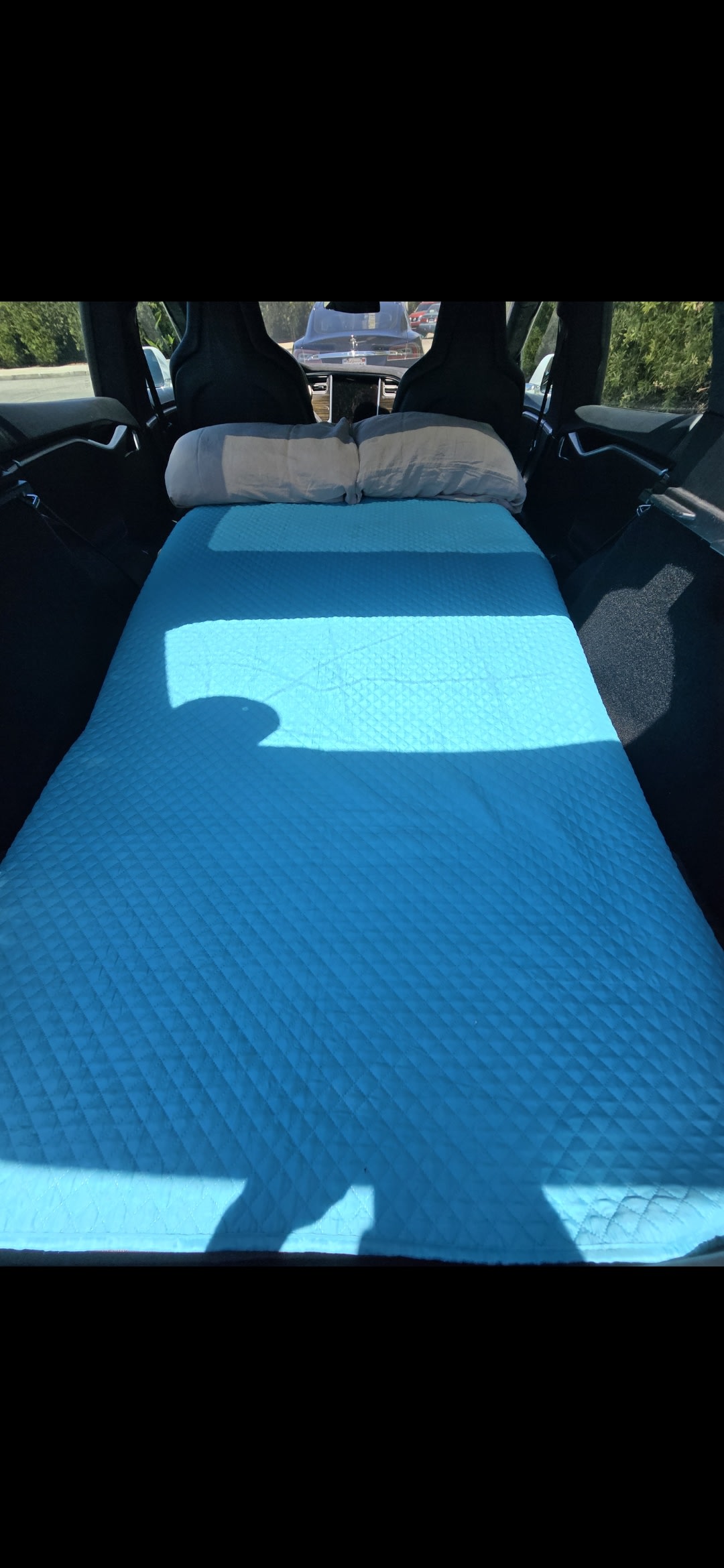 Car's rear transformed into a sleep setup: bright blue quilted mattress & grey pillows visible. Perfect for camping, like an RV rental.. Tesla S 2015