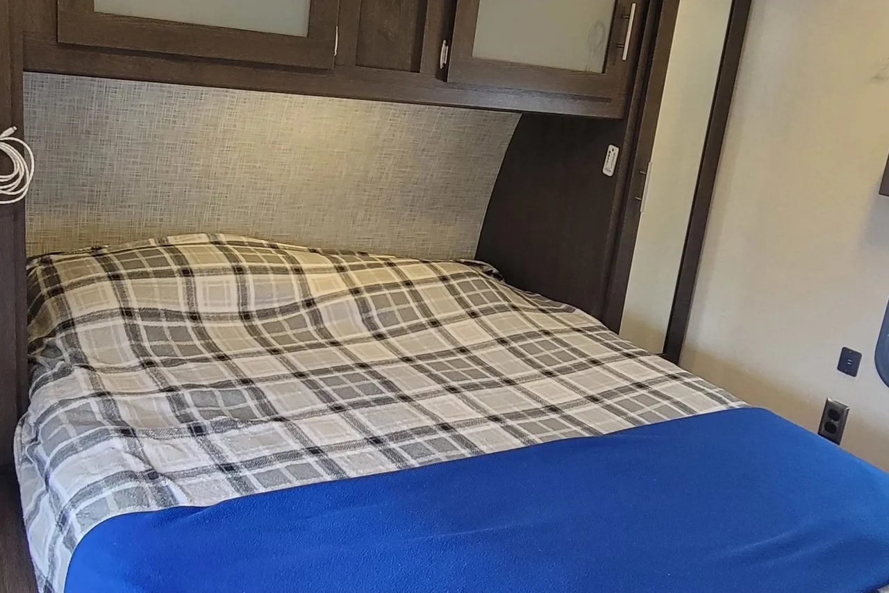 Cozy rv rental bedroom showing a plaid comforter, blue blanket, dark wood overhead cabinets, and a textured headboard.. Forest River Salem 2020