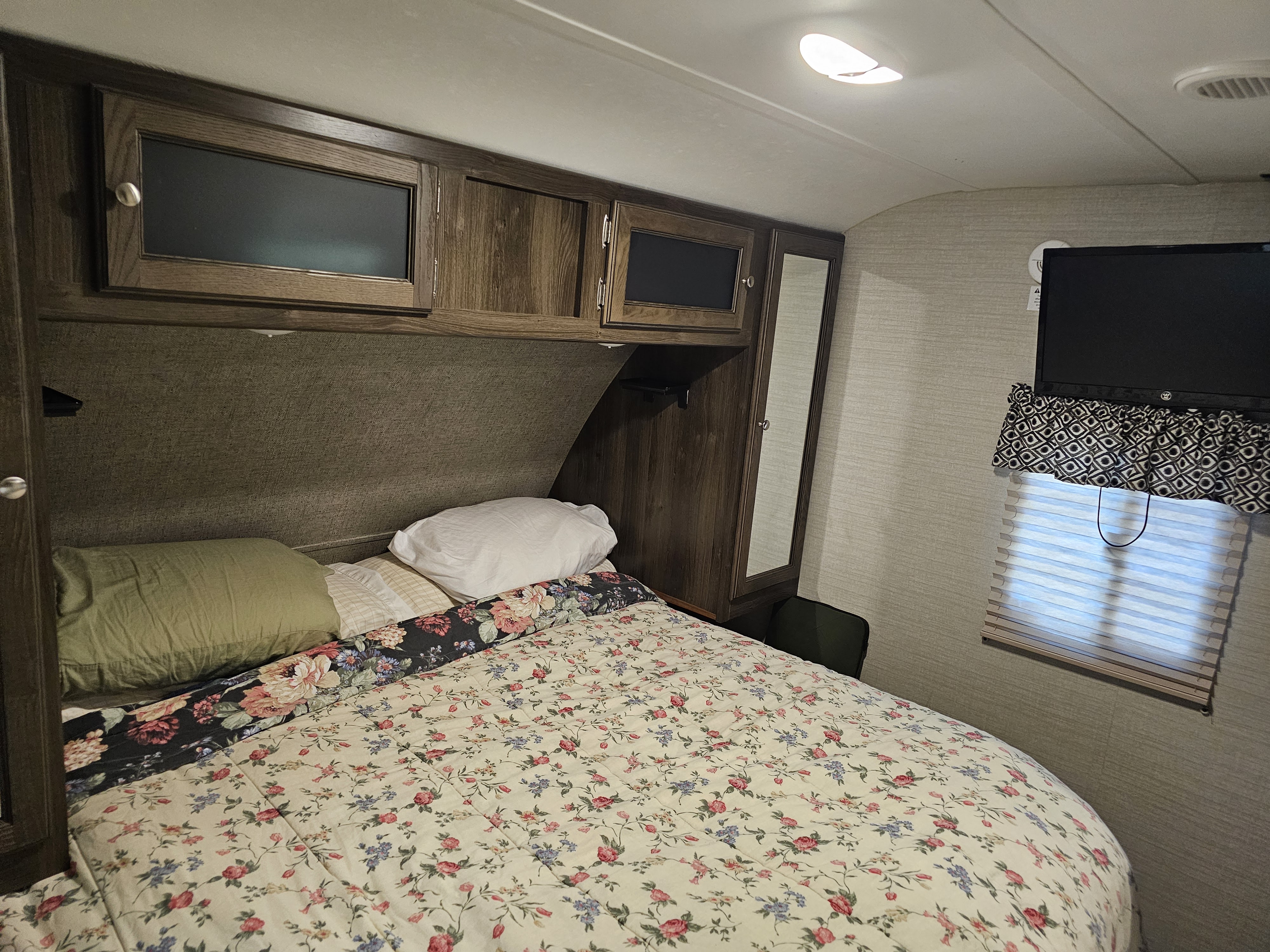 This RV rental's cozy bedroom features a bed with floral bedding, wooden overhead cabinets, a TV, and window with blinds.. Keystone RV Bullet Ultra Lite 2018