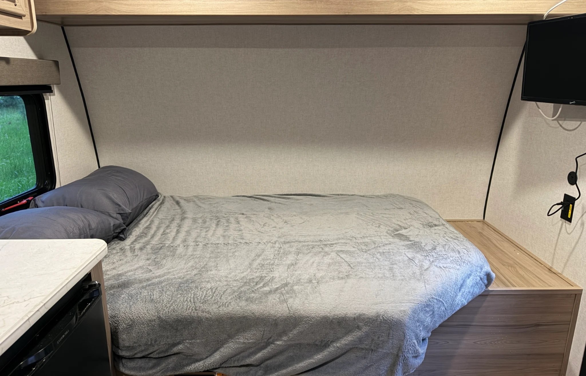 Cozy RV rental bedroom interior with a comfortable bed, gray bedding, a window overlooking greenery, and a mounted TV.. Keystone RV Coleman 2026