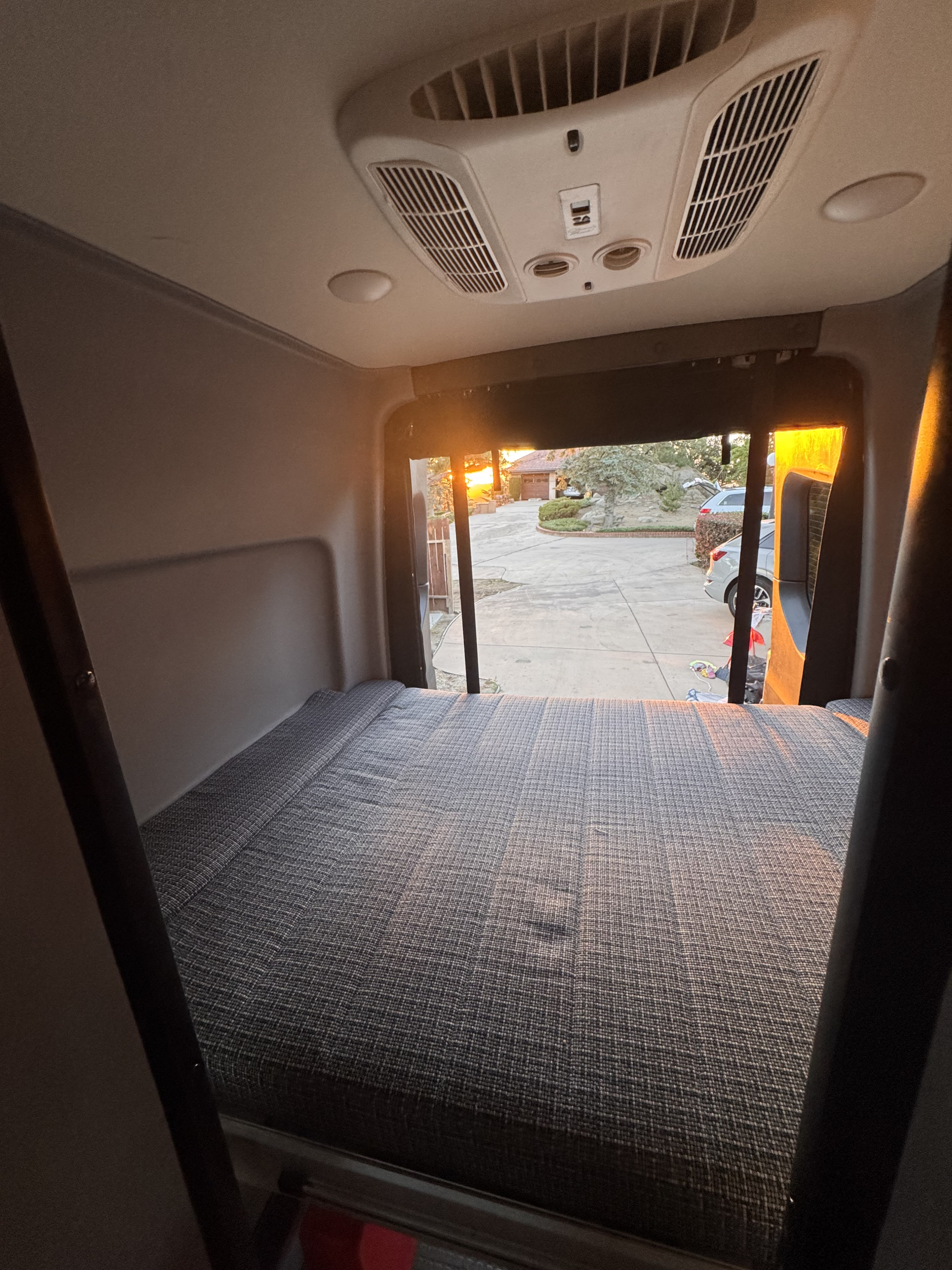Inside an **rv rental** or camper van, a large bed is visible, with open back doors revealing a driveway, house, and a golden sunset.. Winnebago Revel 2022