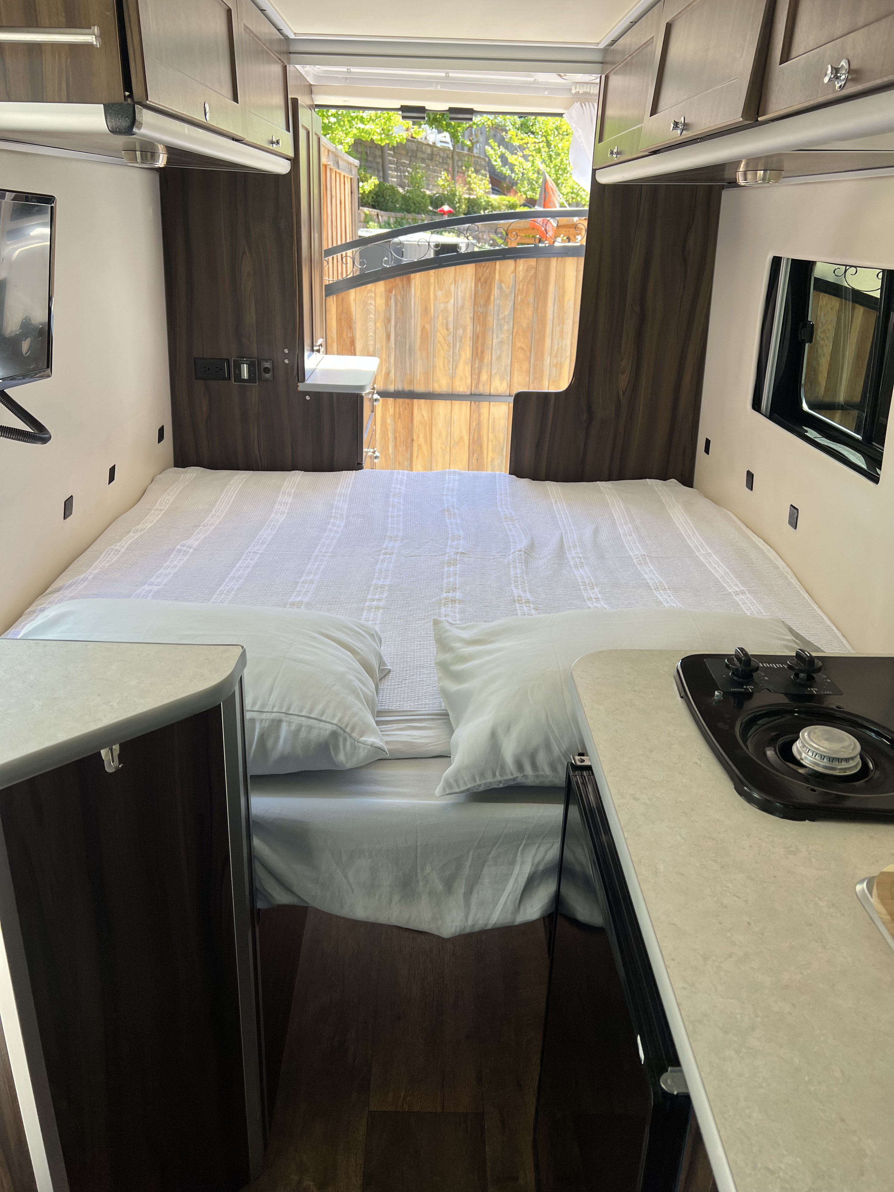 Inside a bright RV rental, showcasing a large bed, compact kitchenette with stovetop, TV, and open rear door revealing an outdoor scene.. Hymer Carado Banff 2017