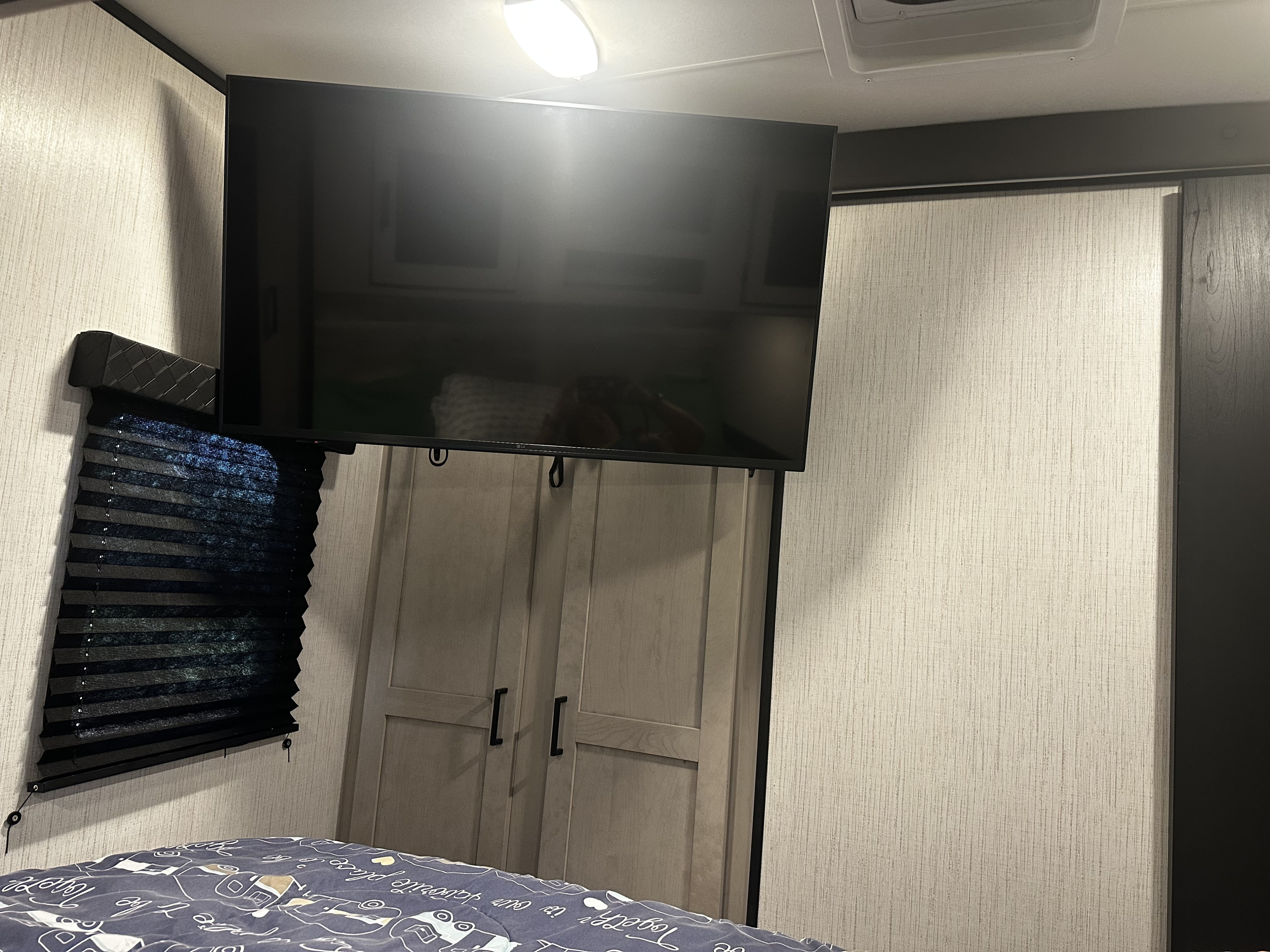 Inside an RV rental, a mounted TV overlooks a closet & bed. A dark window blind is on the left wall of this compact space.. Heartland RVs Mallard 2022