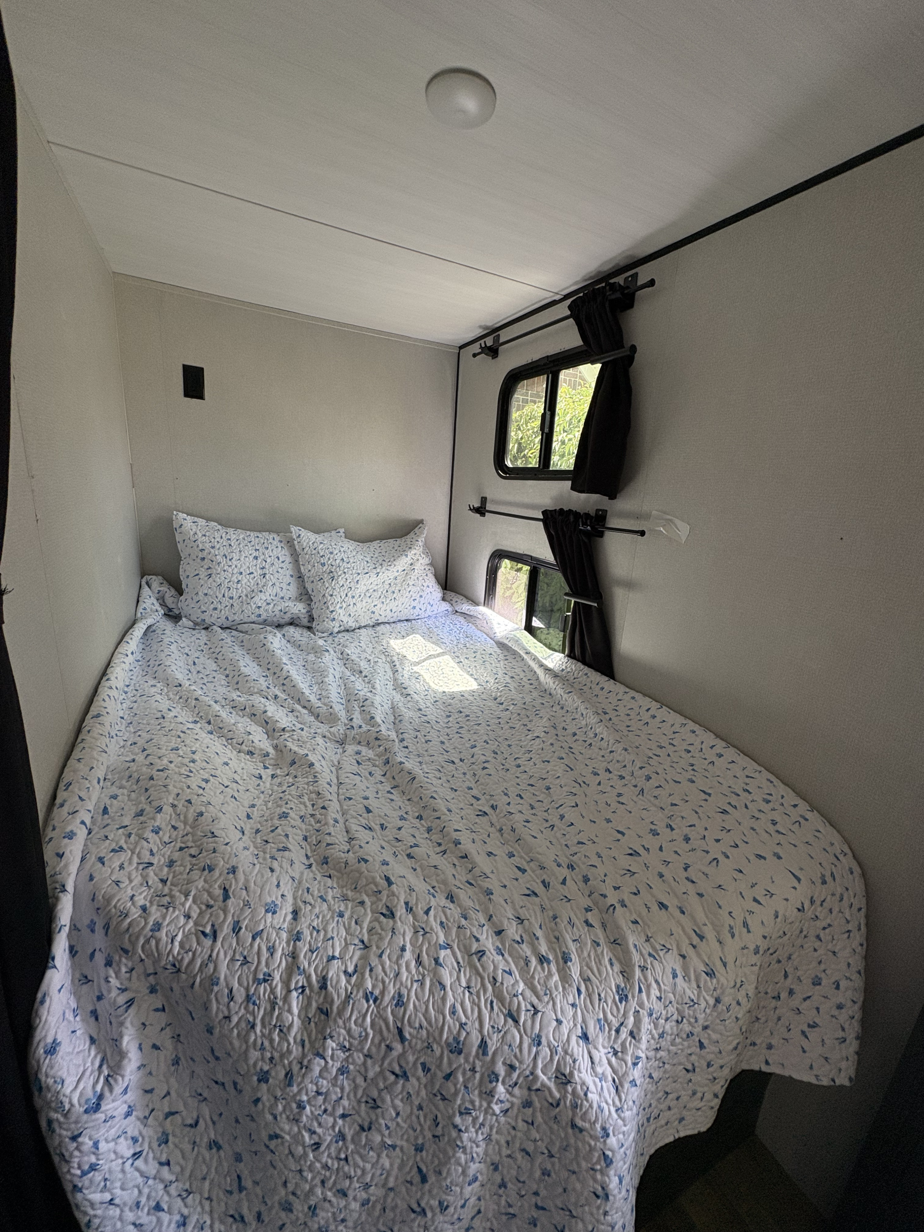 Cozy RV rental bedroom with a floral quilt and pillows, featuring small windows with black curtains and sunlight.. Keystone RV Hideout 2022