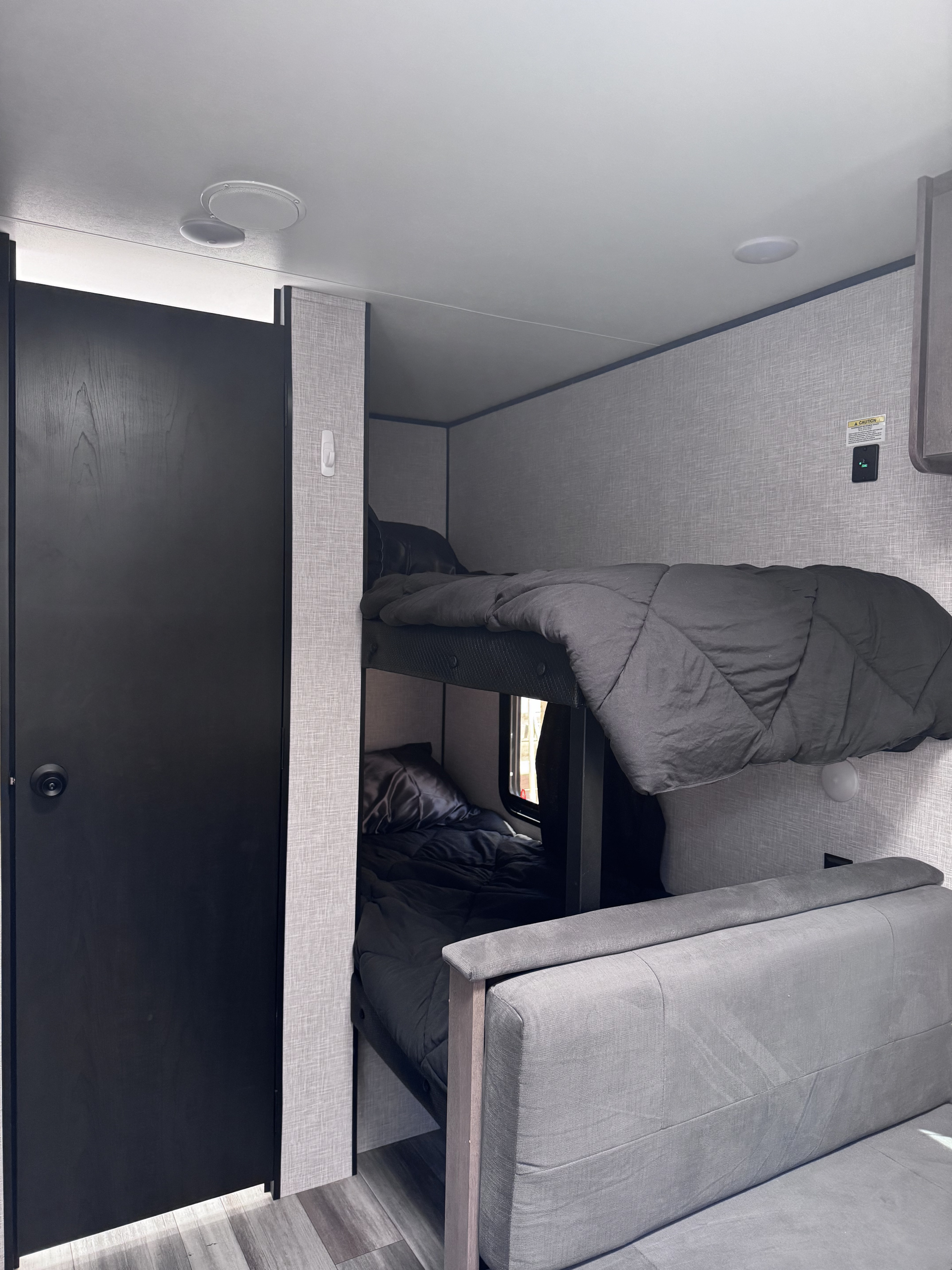 RV rental interior featuring cozy bunk beds with dark bedding, a gray couch, and a black door on the left.. Jayco Jay Flight 2025