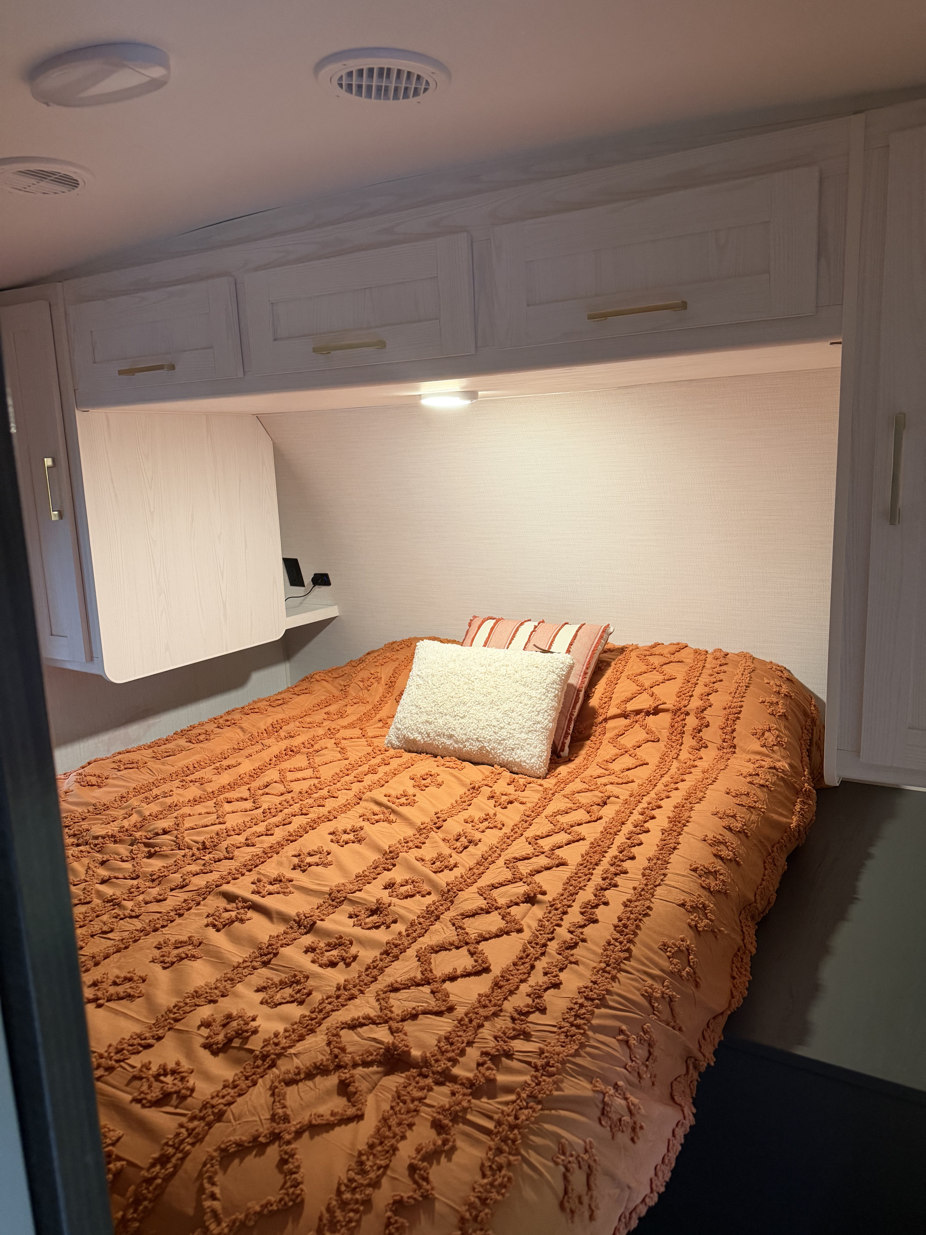 Cozy RV rental bedroom featuring an orange textured comforter, decorative pillows, light overhead cabinets, and integrated lighting.. Heartland RVs Eddie Bauer 2024