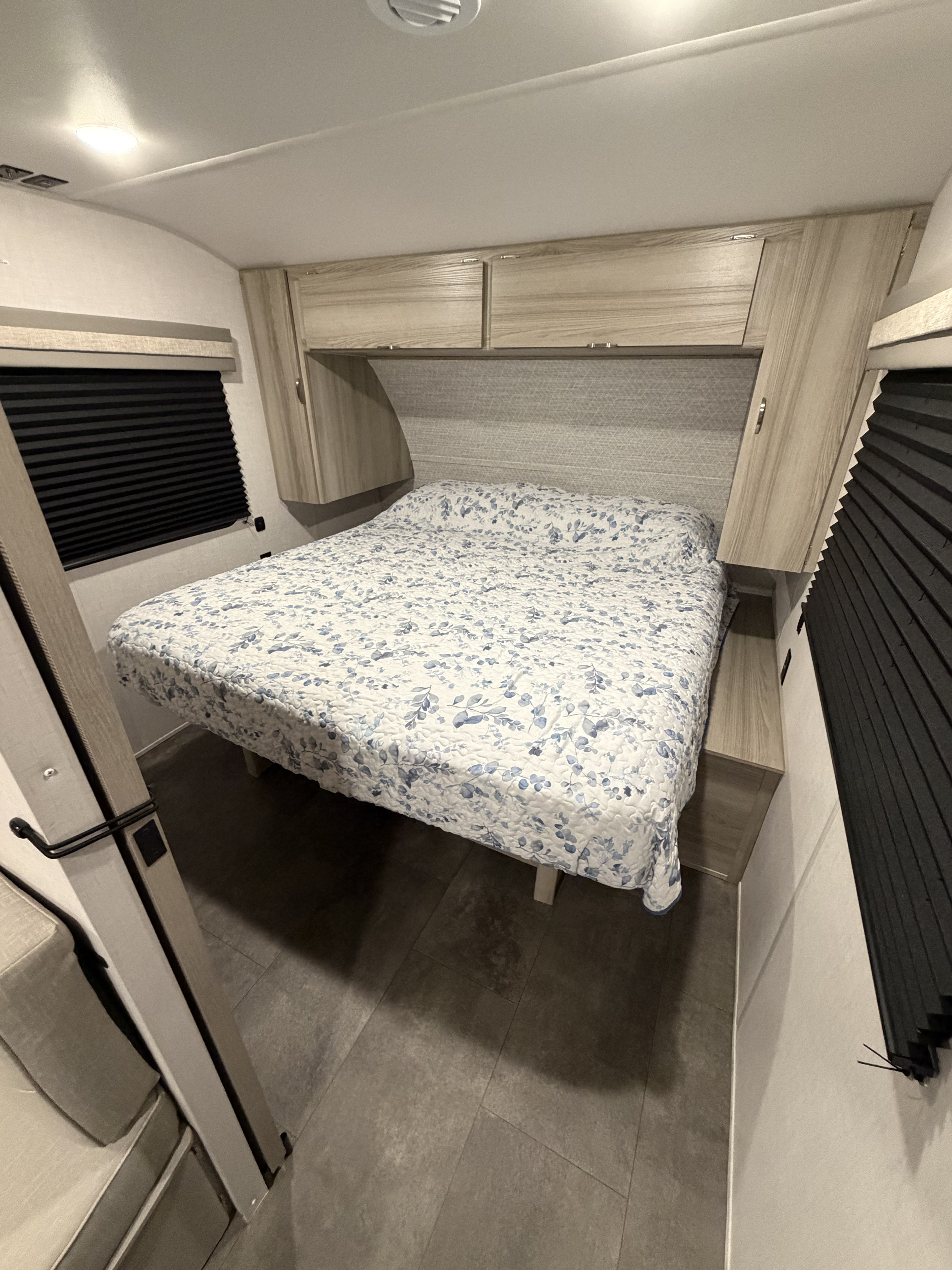 Cozy RV rental bedroom with a floral bedspread, light wood overhead cabinets, dark pleated blinds, and tiled floor.. Forest River Wildwood FSX 2024
