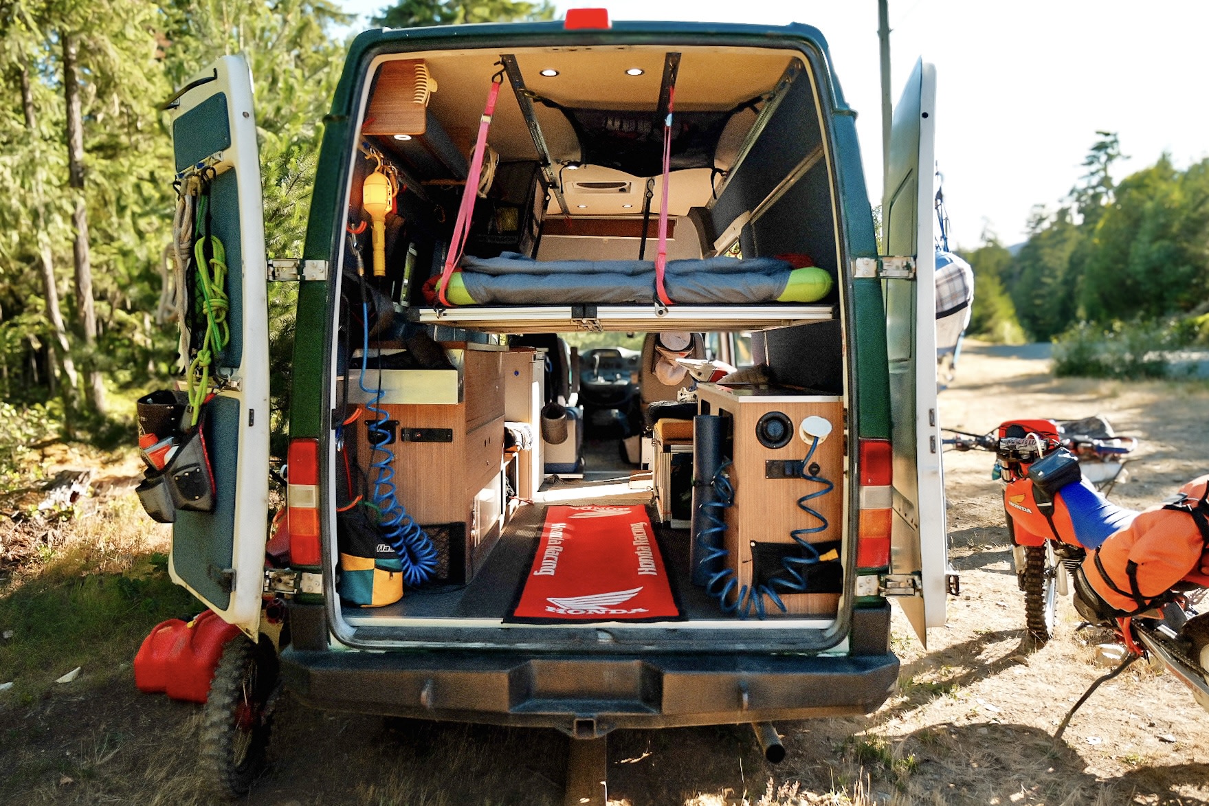 This RV rental camper van showcases its custom interior, featuring a bed, storage, and gear, with a dirt bike nearby.. Mercedes-Benz Sprinter 2006
