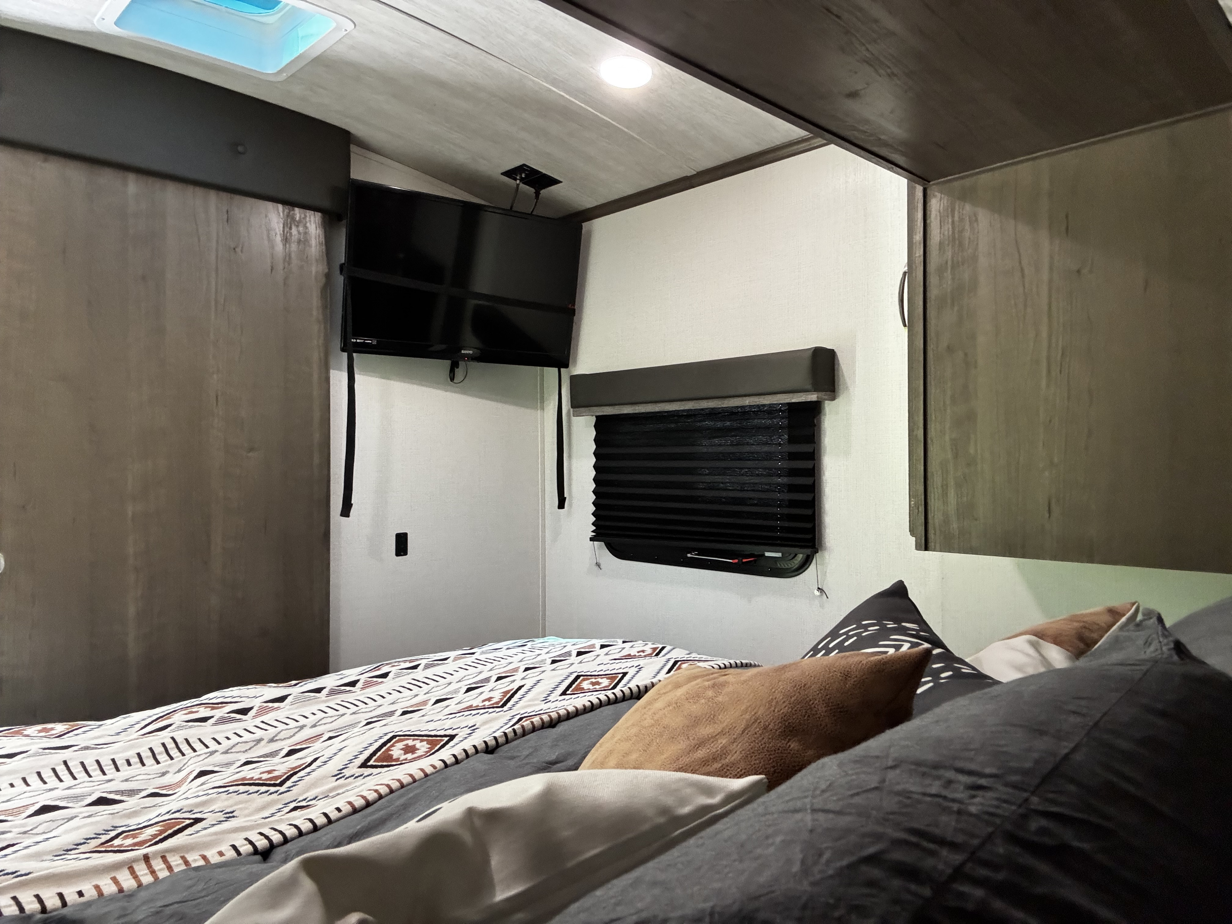 Inside an RV rental bedroom featuring a mounted TV, pleated window blind, and comfy bed with tribal-patterned blanket.. Keystone RV Cougar 2021
