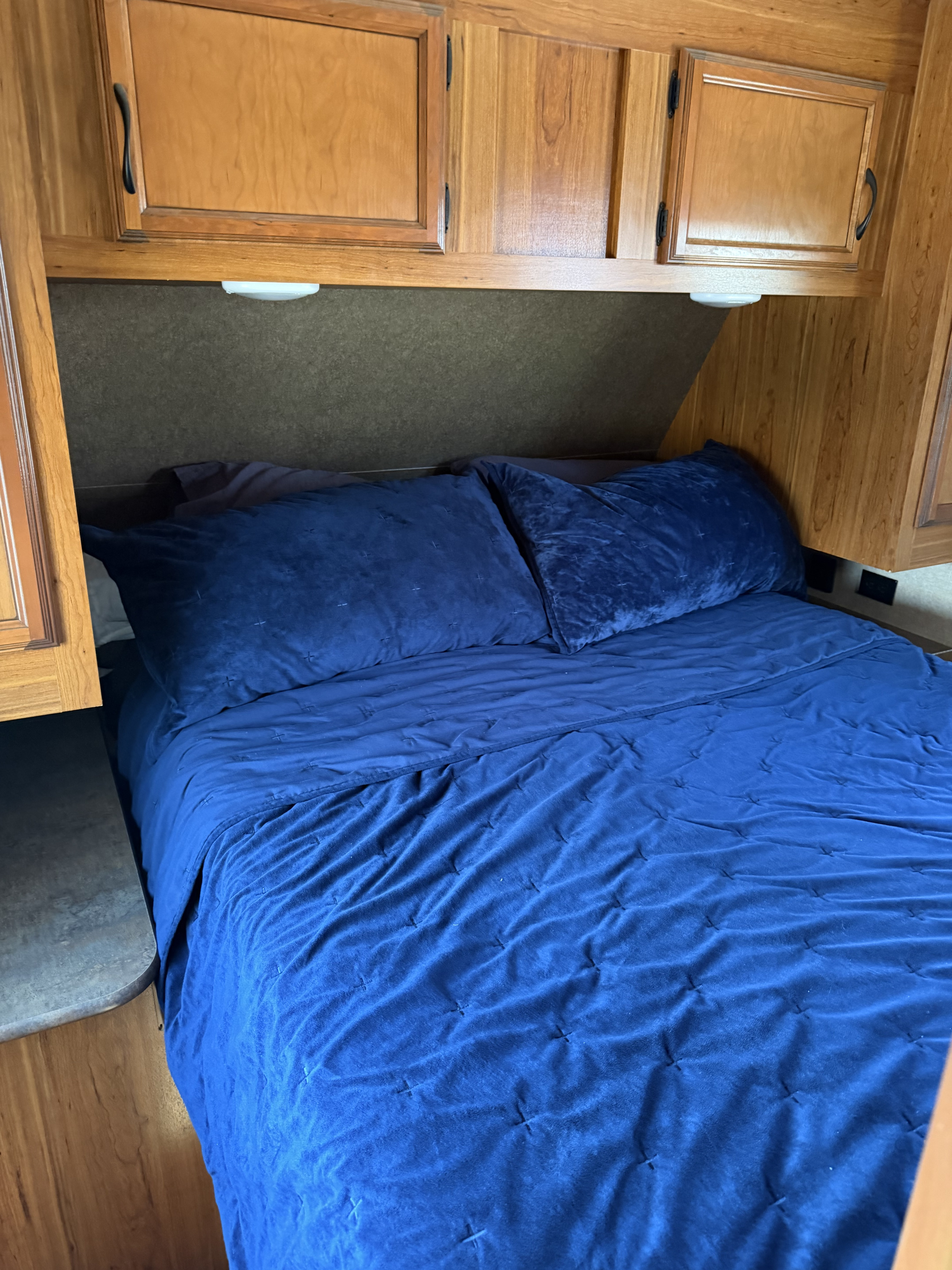 A cozy blue bed with quilted bedding inside an RV rental, featuring warm wooden overhead cabinets and built-in lighting.. Starcraft Autumn Ridge 2015