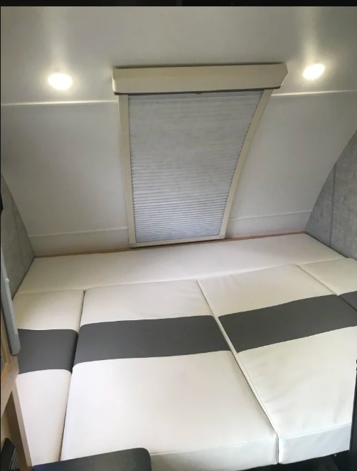Comfortable RV rental bed with white and gray cushions, overhead lighting, and a window blind, perfect for a cozy trip.. Xtreme Outdoors Xtreme Outdoors Trailer 2021