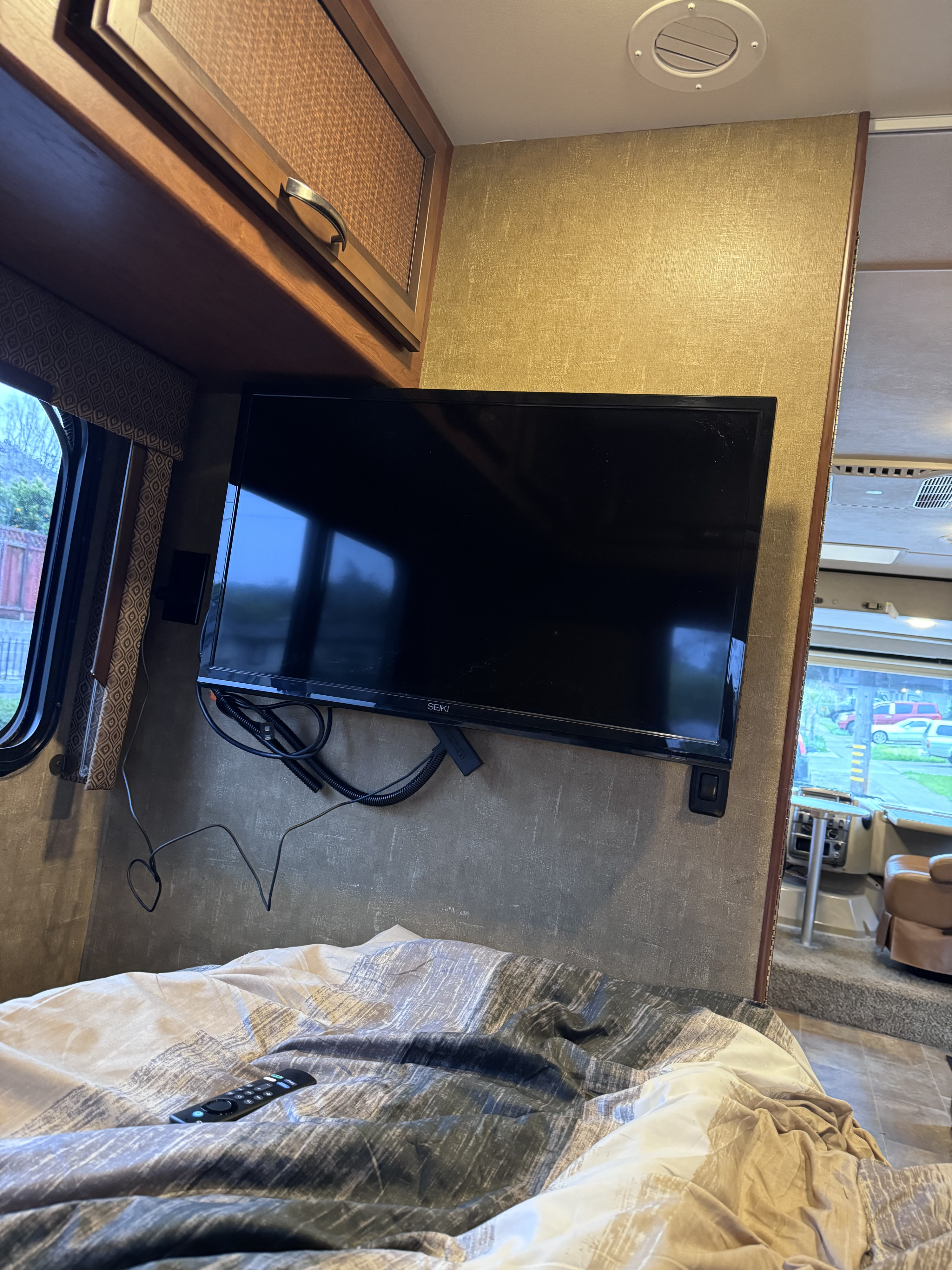 RV rental bedroom with a wall-mounted TV, remote on a patterned comforter, overhead cabinet, and a window view.. Thor Axis 2015