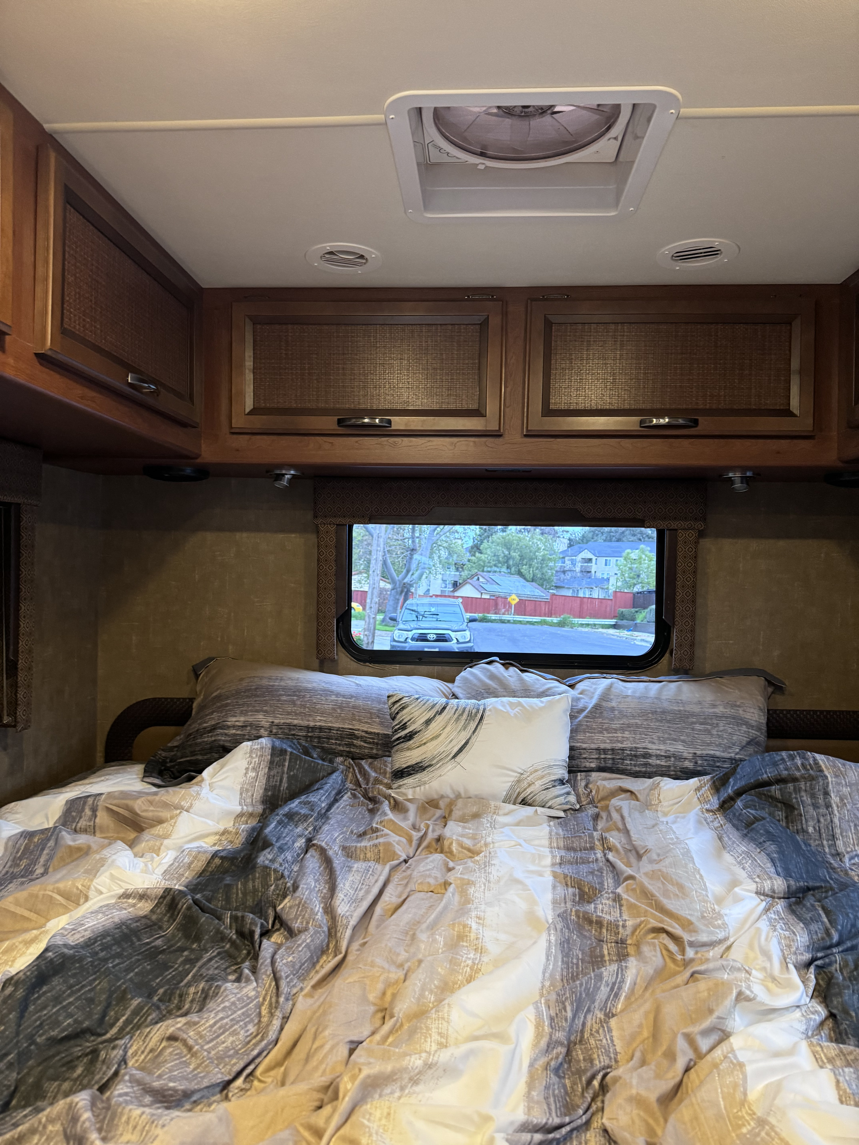 RV rental bedroom with patterned bedding, pillows, wooden overhead cabinets, a ceiling vent, and a window view.. Thor Axis 2015