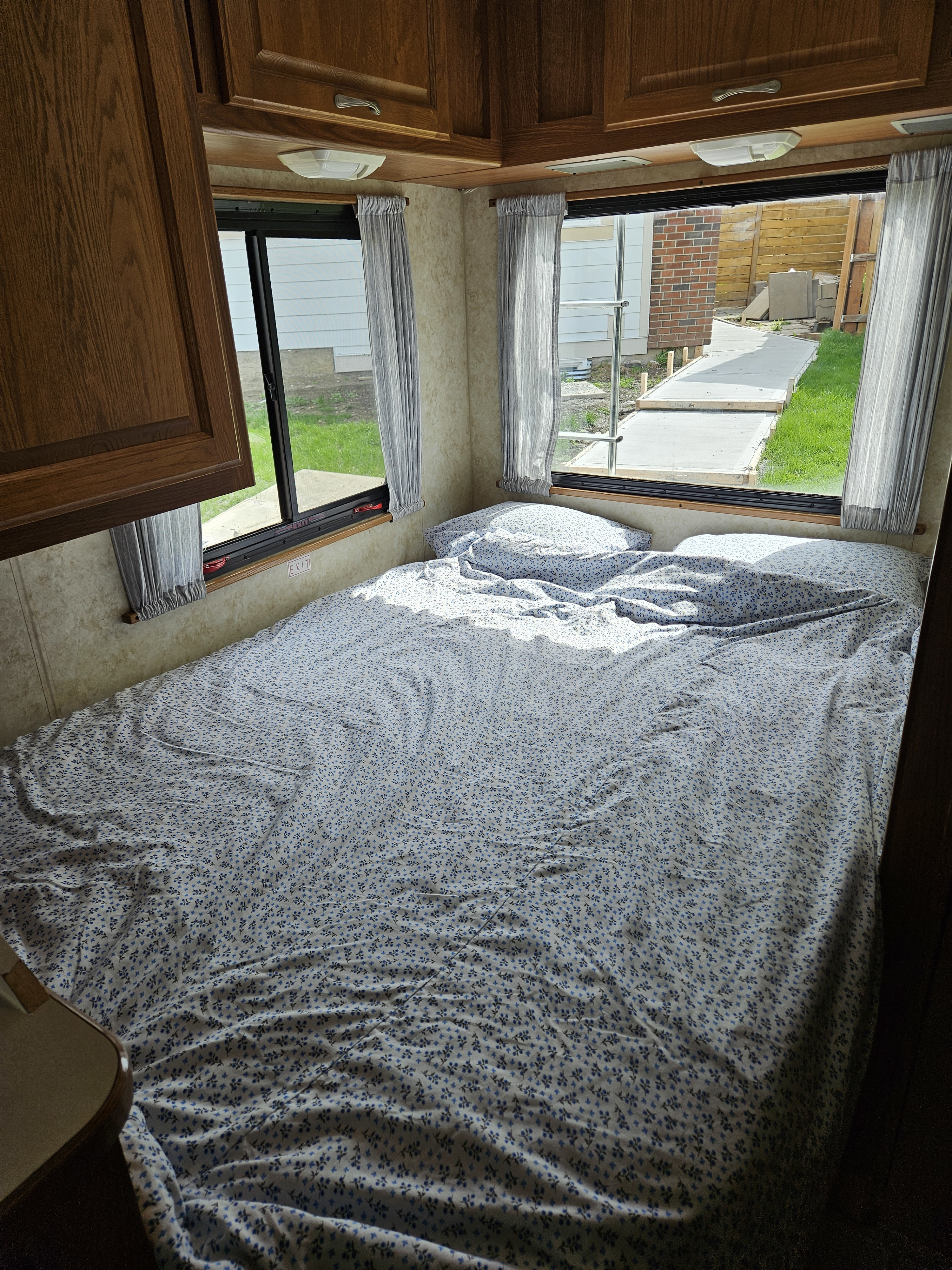 Inside an RV rental: a bright, cozy bedroom with blue floral bedding, wooden cabinets, and windows showing a sunny outdoor path.. Ford Majestic 2004