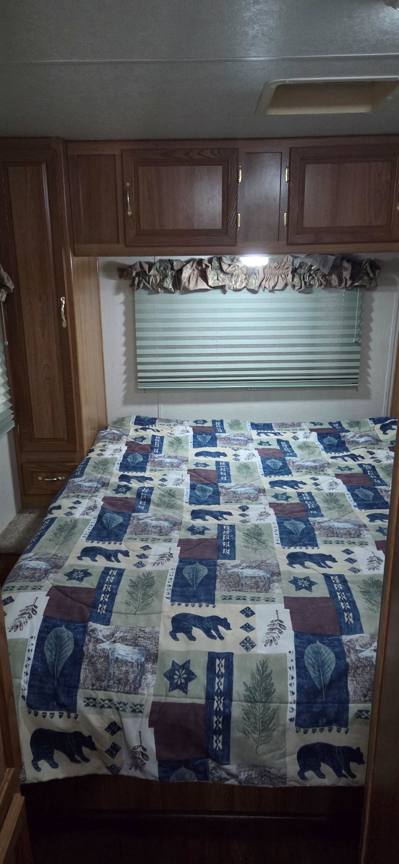 Cozy rv rental bedroom features a nature-themed comforter, wooden cabinets, and a window with blinds.. Coachmen Leprechaun 2009