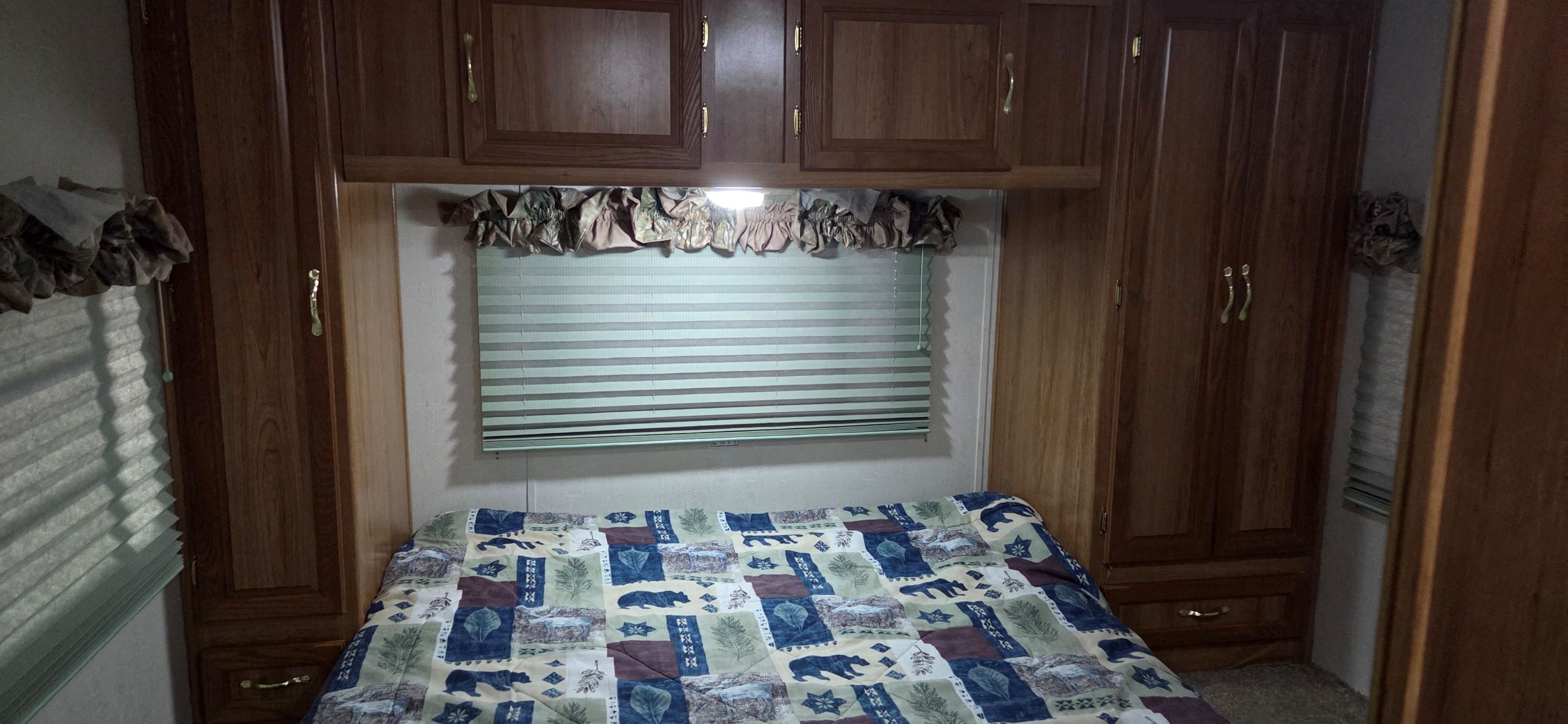 RV rental bedroom featuring wood cabinetry, a window with blinds, and a nature-patterned bedspread. Compact and functional space.. Coachmen Leprechaun 2009